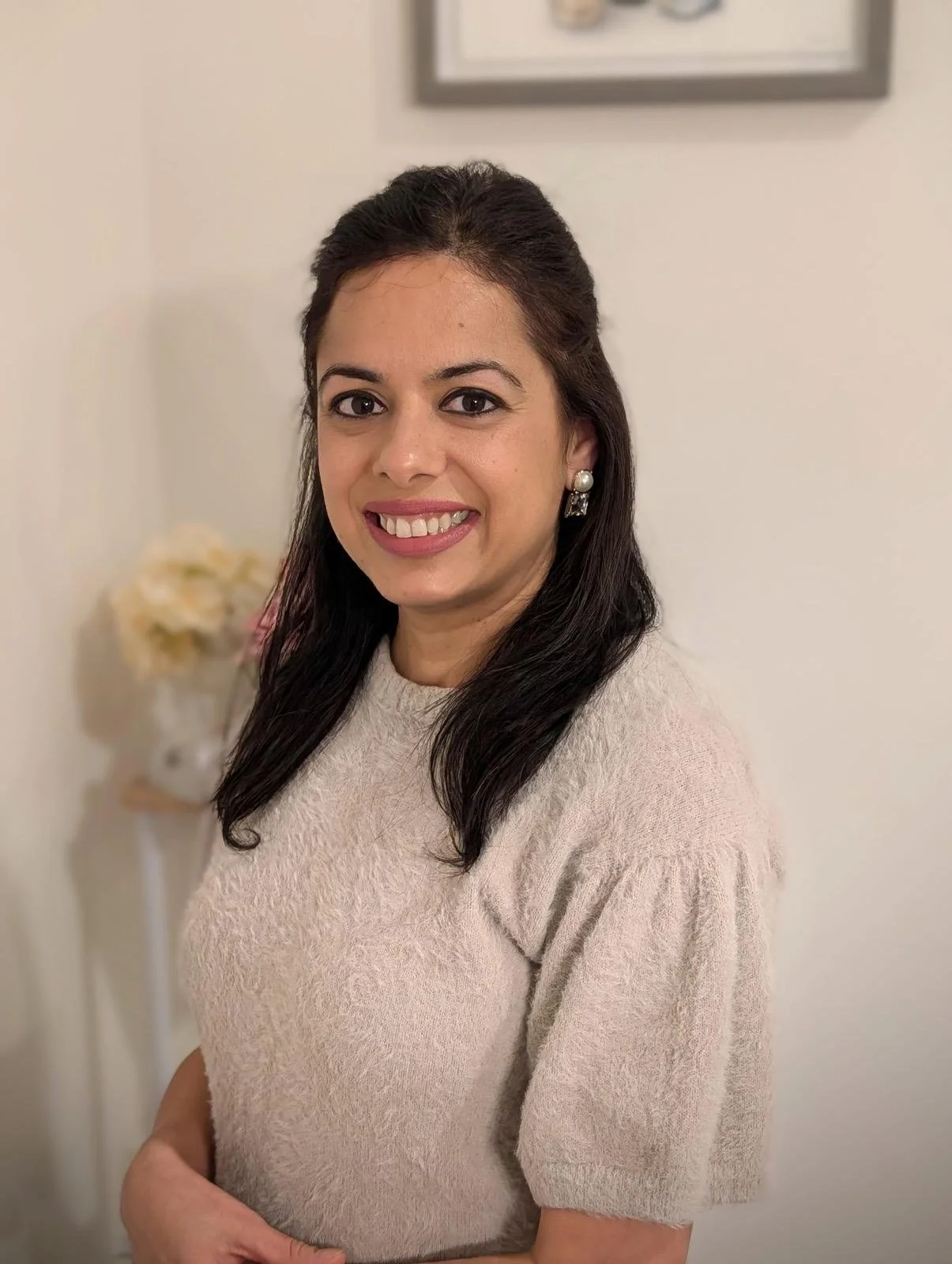 A woman with dark hair, wearing a light beige sweater and pearl earrings, smiling at the camera in a room with beige walls and a framed picture and flowers in the background.