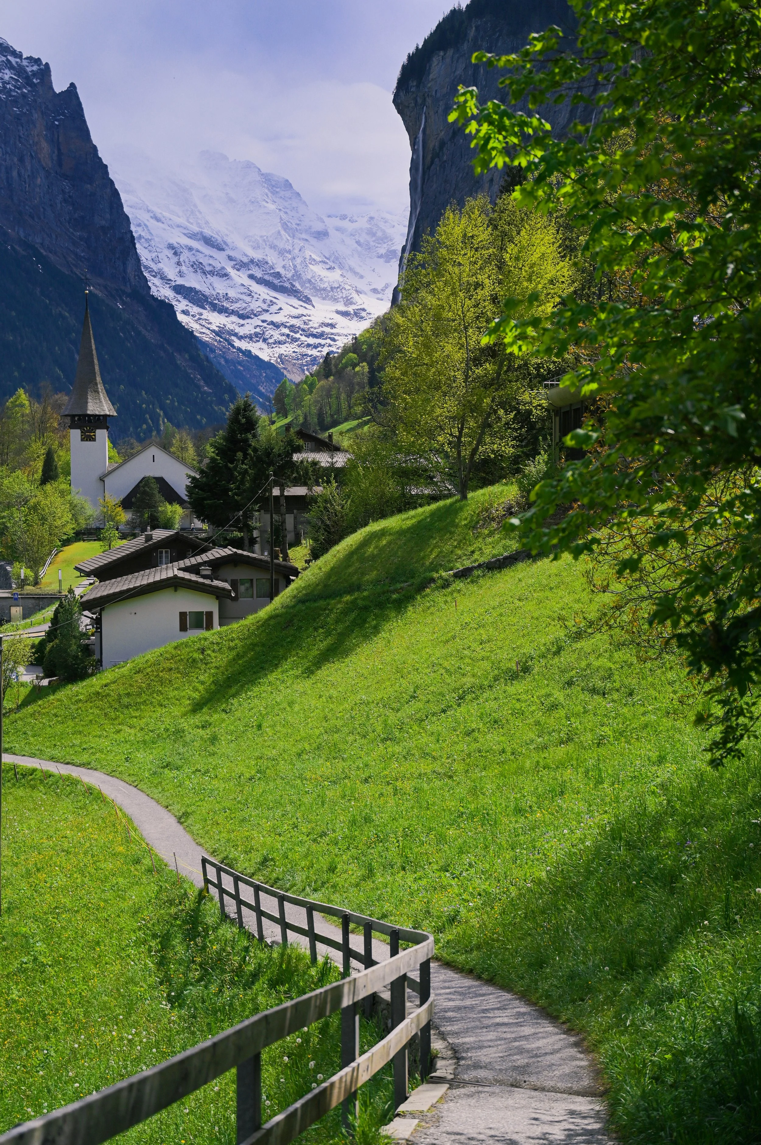 A winding path through a lush green hillside in a mountainous region with snow-capped peaks in the background. There are several houses and a church with a tall steeple, and trees scattered across the landscape.