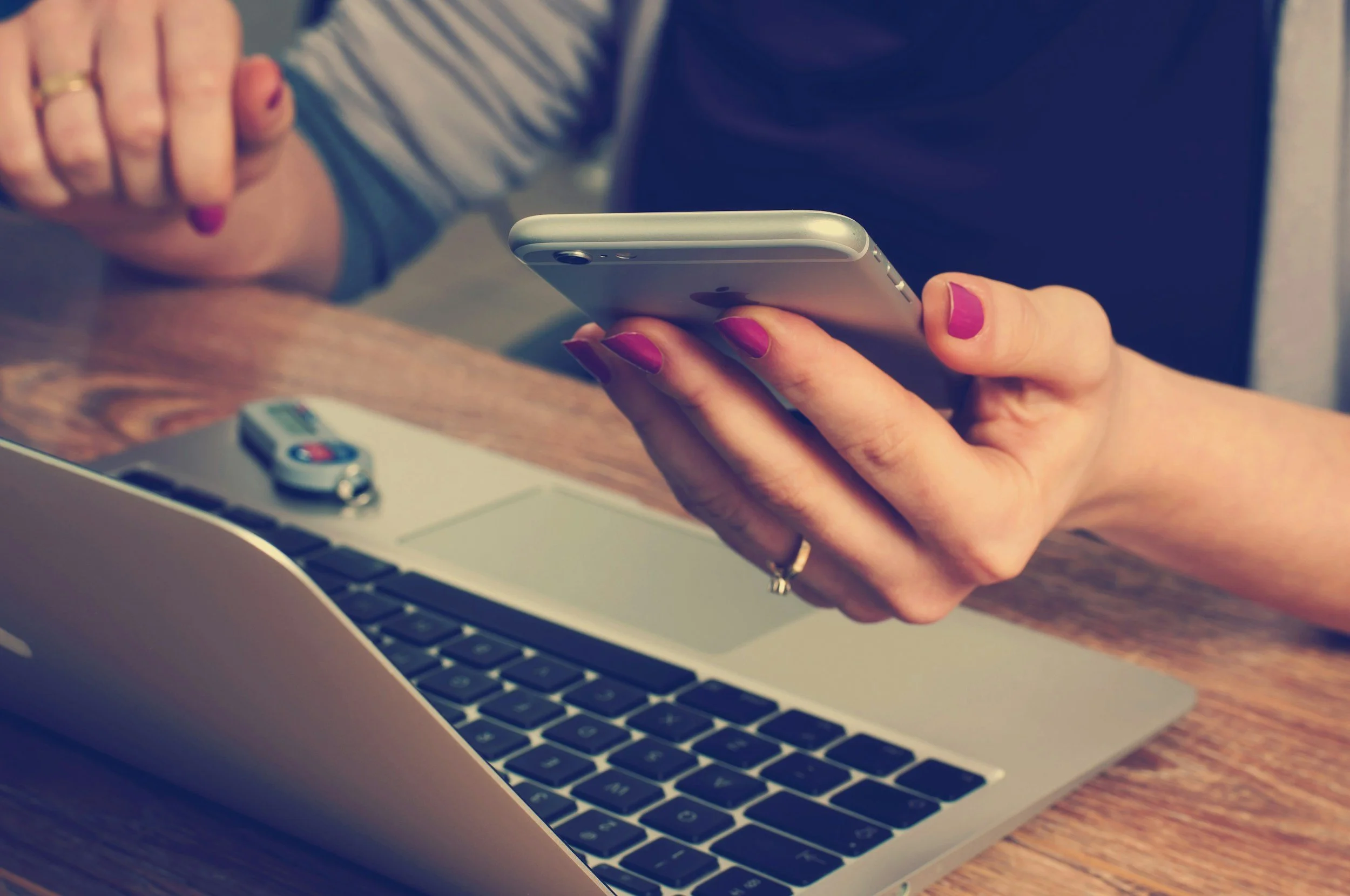 A person holding a smartphone with a laptop and car key on the wooden table.