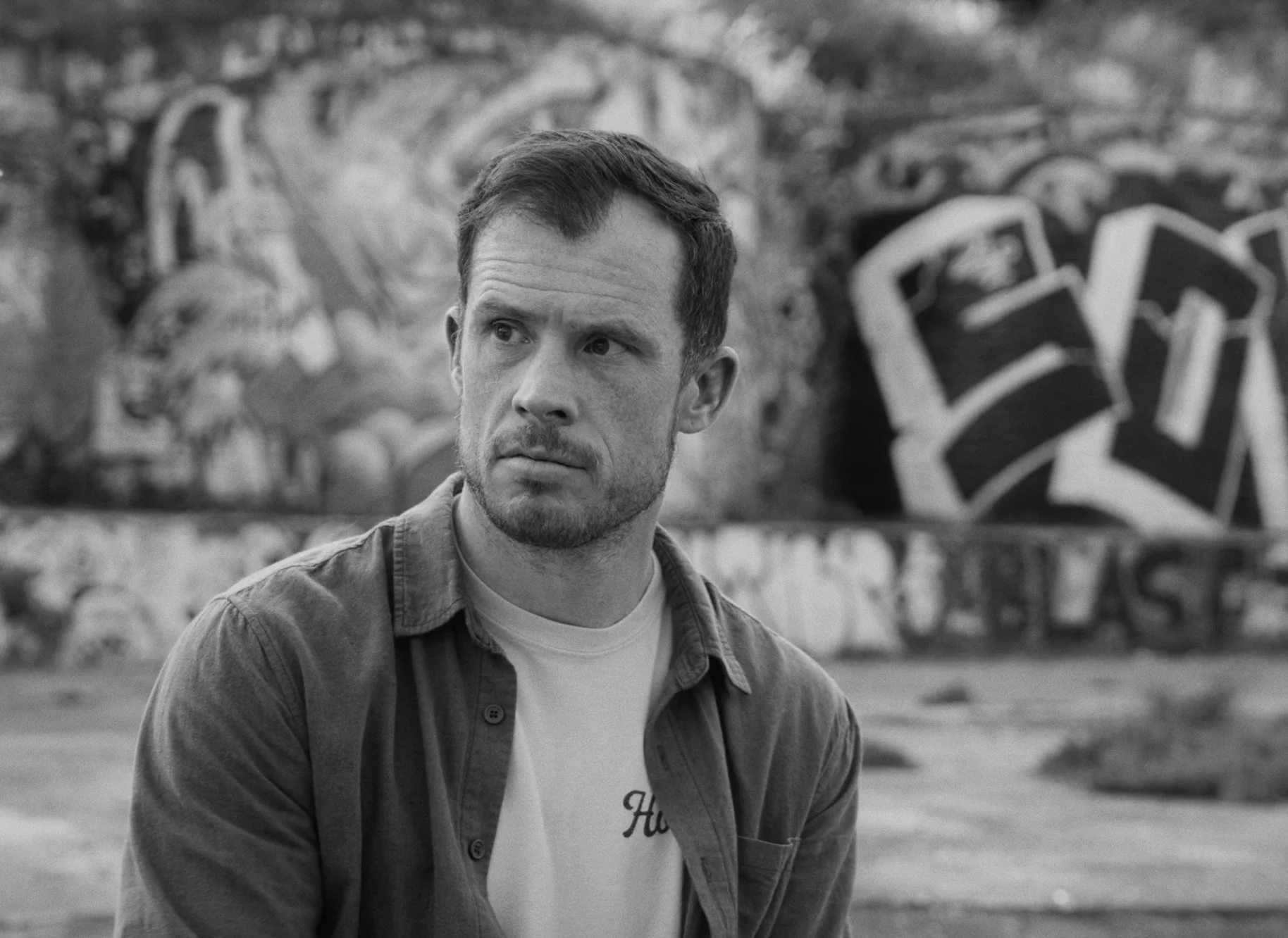 A man with short hair and a light beard looking off to the side, wearing a light shirt and a jacket, standing outdoors with graffiti art in the background.