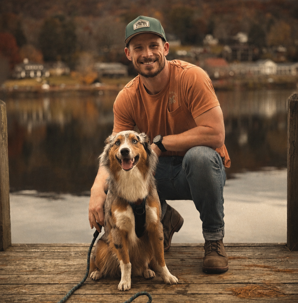 A man crouching on a wooden dock, smiling, with an adorable Australian Shepherd dog sitting next to him, near a calm lake with autumn-colored trees and houses in the background.