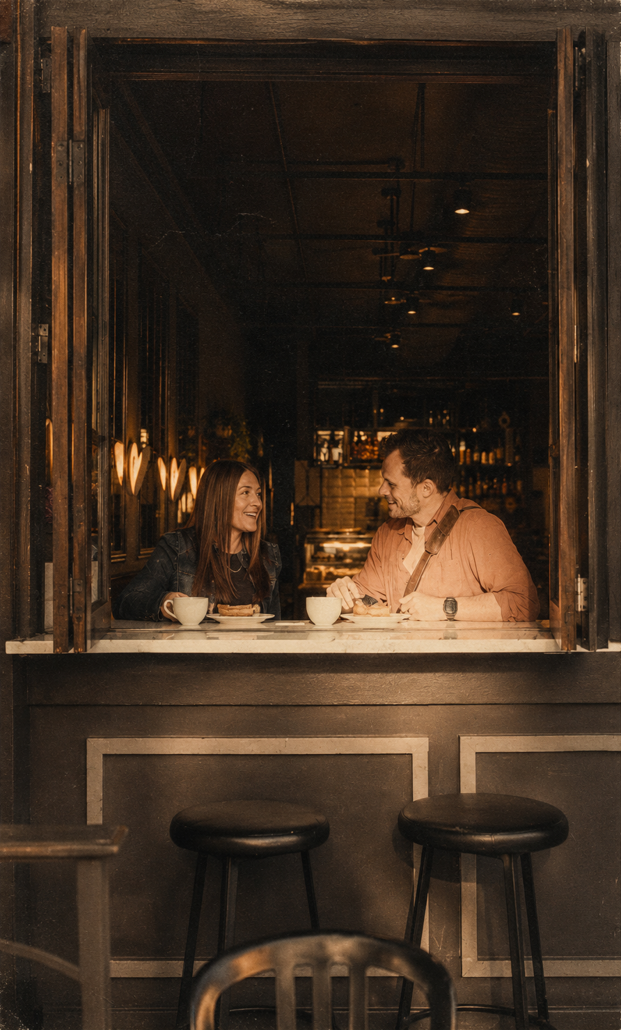 A man and woman sitting at a counter in a dimly lit cafe or restaurant, enjoying coffee and conversation, with a cozy atmosphere.