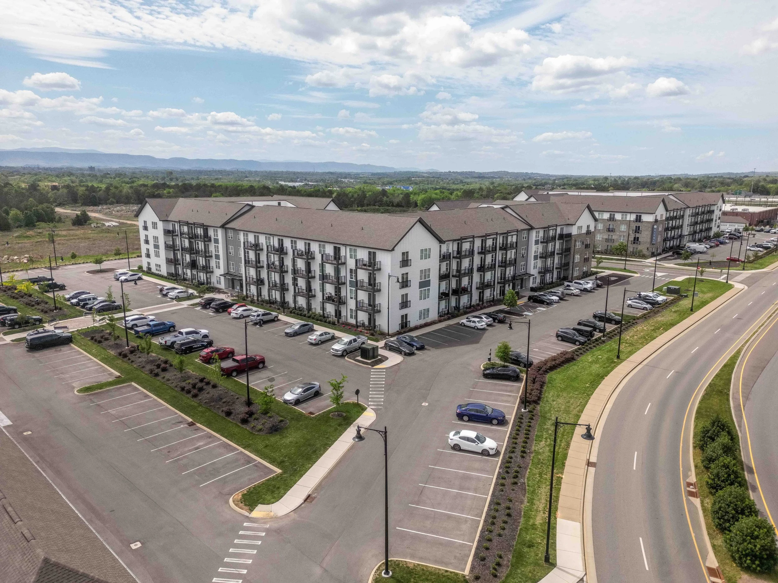 An aerial view of a residential apartment complex with multiple buildings, parking lots filled with cars, landscaped areas, and a road. In the background, there are lush green trees, open land, and mountains under a partly cloudy sky.