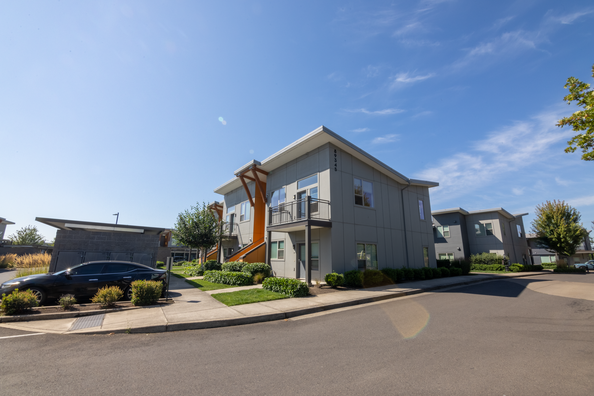 Modern residential apartment complex with multiple units, green shrubbery, a parked black car, and a clear blue sky.