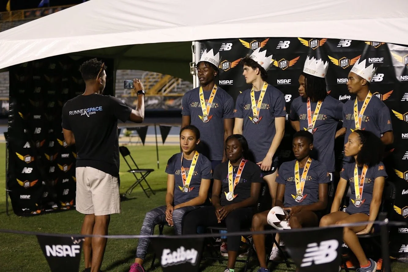 A group of young athletes, wearing medals and crown-like hats, pose for a photo at an awards ceremony in a sports stadium, with a man taking their picture.