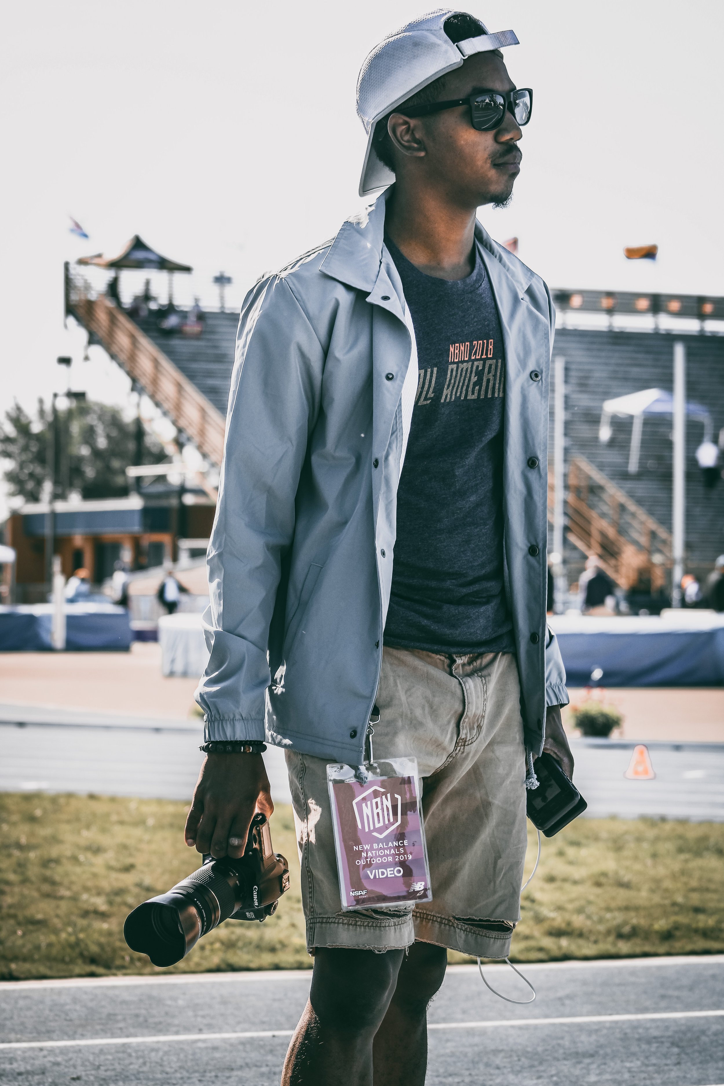A young man wearing a white cap backwards, black sunglasses, a dark t-shirt, and a light jacket, holding a camera in one hand and a phone in the other, standing outdoors near a track with a crowd in the background during daytime.