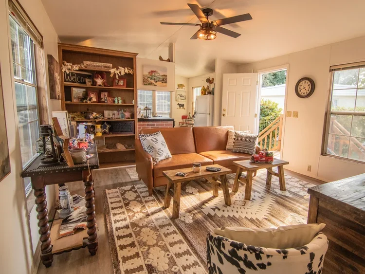 Living room with a brown leather couch, wooden coffee tables, a patterned armchair, and a large window. There is a wooden bookshelf with decorations and books, a ceiling fan, and a white door leading outside. Sunlight is streaming in, and various decorative items and furniture are visible.