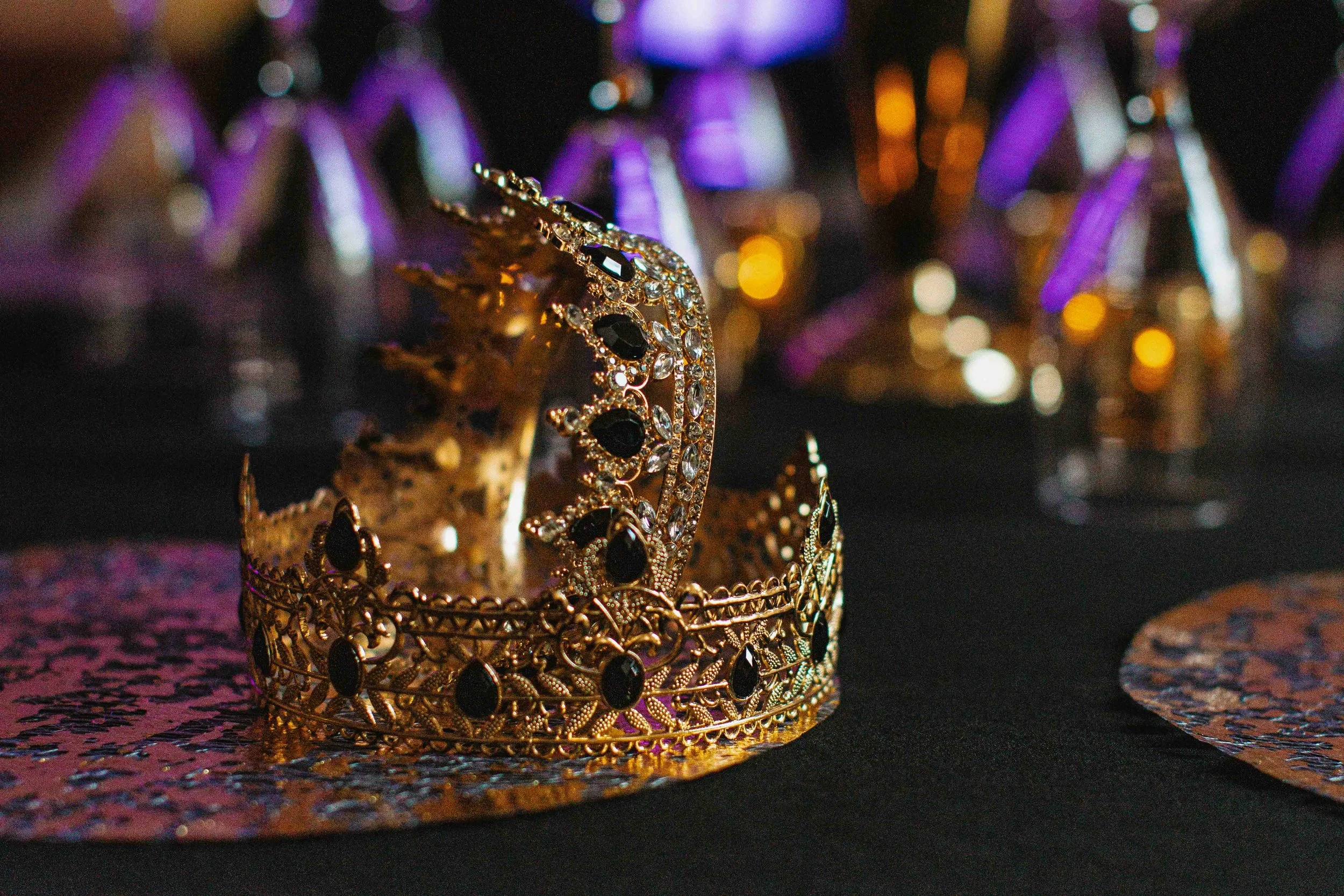 A gold crown decorated with black and clear gemstones is placed on a surface with a shiny, colorful pattern. The background is blurred with purple and gold bokeh lights.