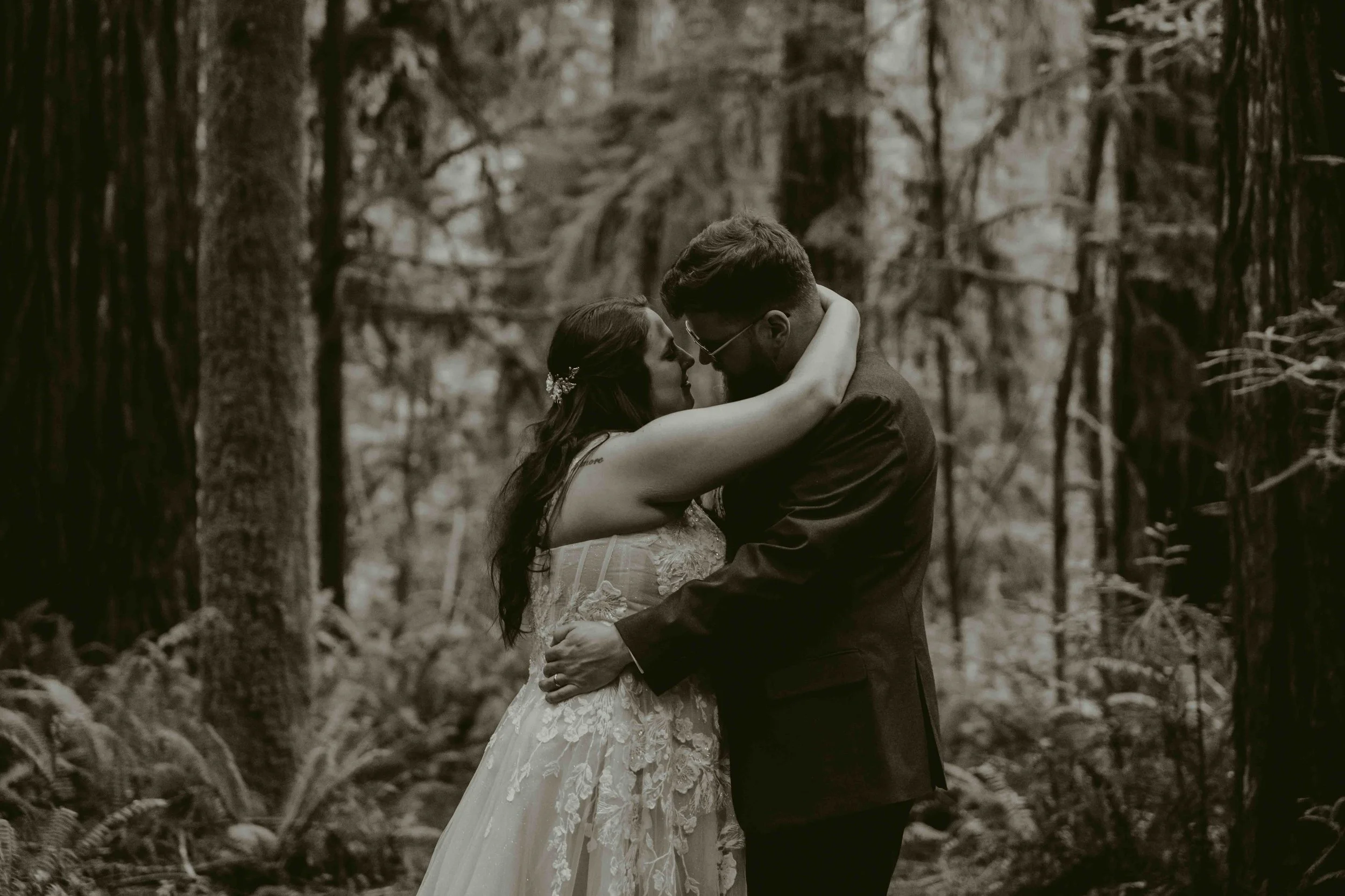 A couple embracing in a forest, with the woman wearing a wedding dress and the man in a suit, in a black and white photo.