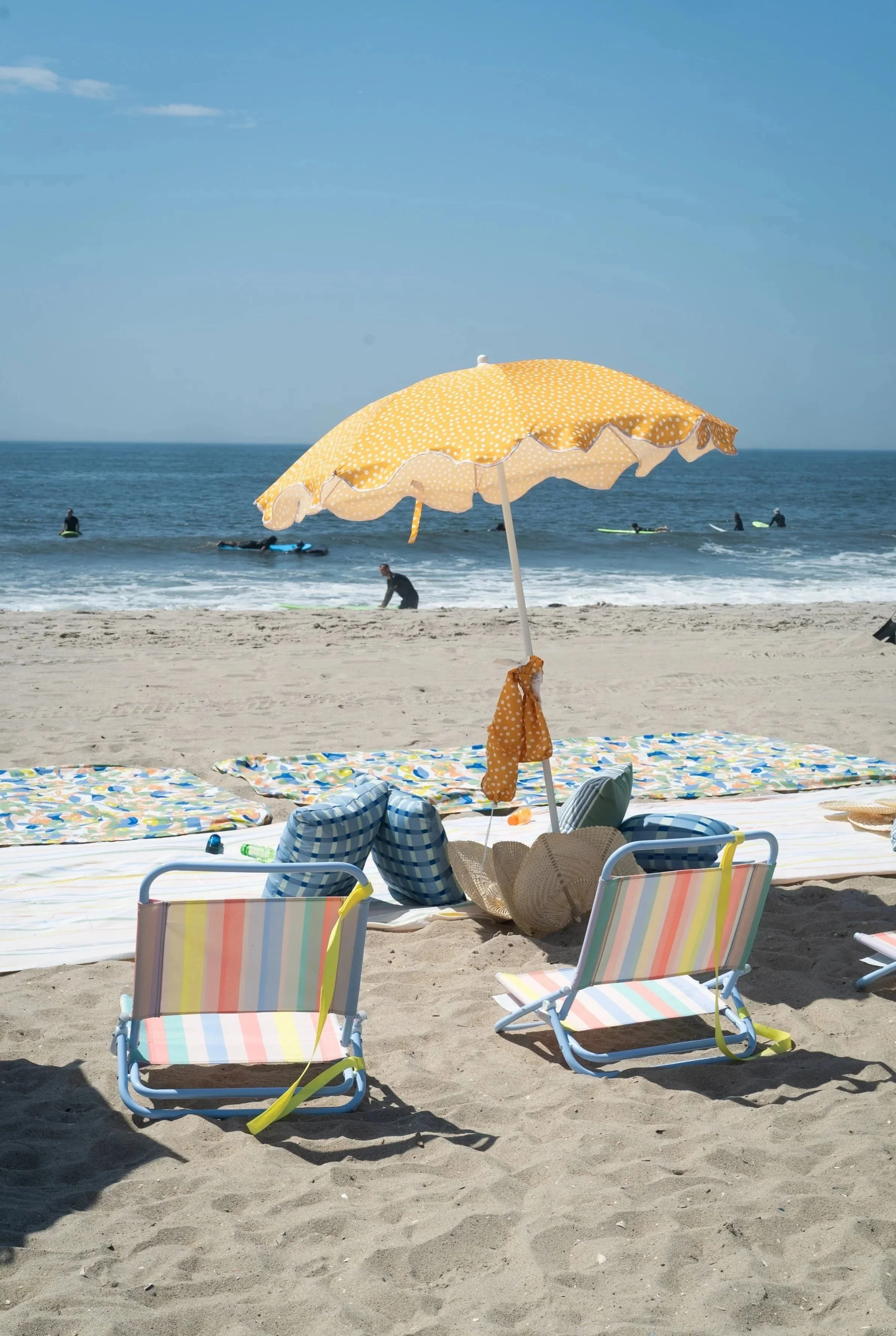 Beach scene during Elsewhere Retreats' Rockaway Reset at Rockaway Beach NY with a yellow polka dot umbrella, two striped beach chairs, and towels on the sand. In the background, people are surfing and swimming in the ocean under a blue sky.