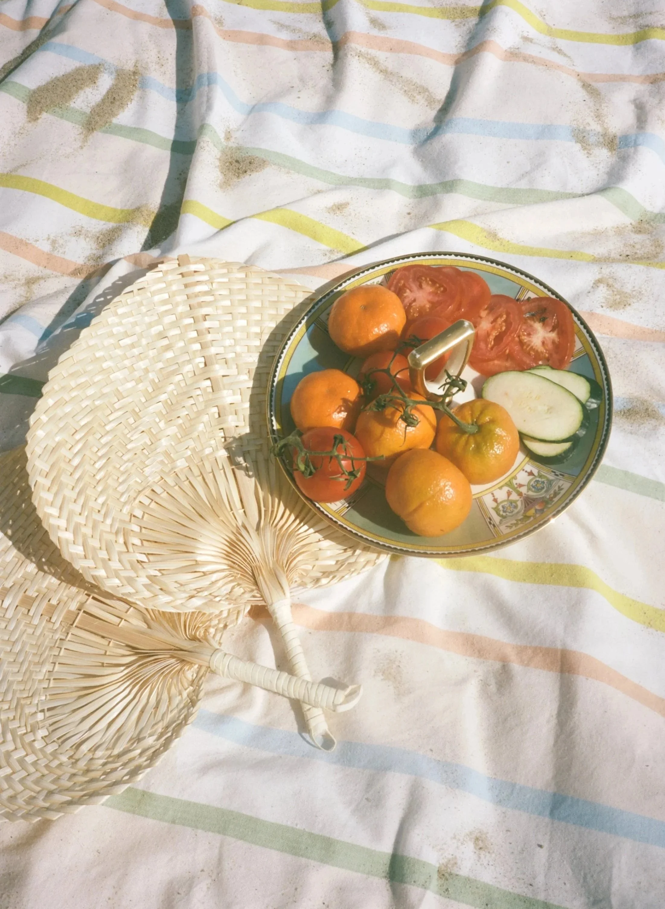 A healthy, delicious, and colorful lunch during Elsewhere Retreats' Rockaway Reset at Rockaway Beach NY featuring sliced tomatoes, cucumber slices, and small orange tomatoes, placed on patterned beach sheet with a woven fan and a basket nearby.