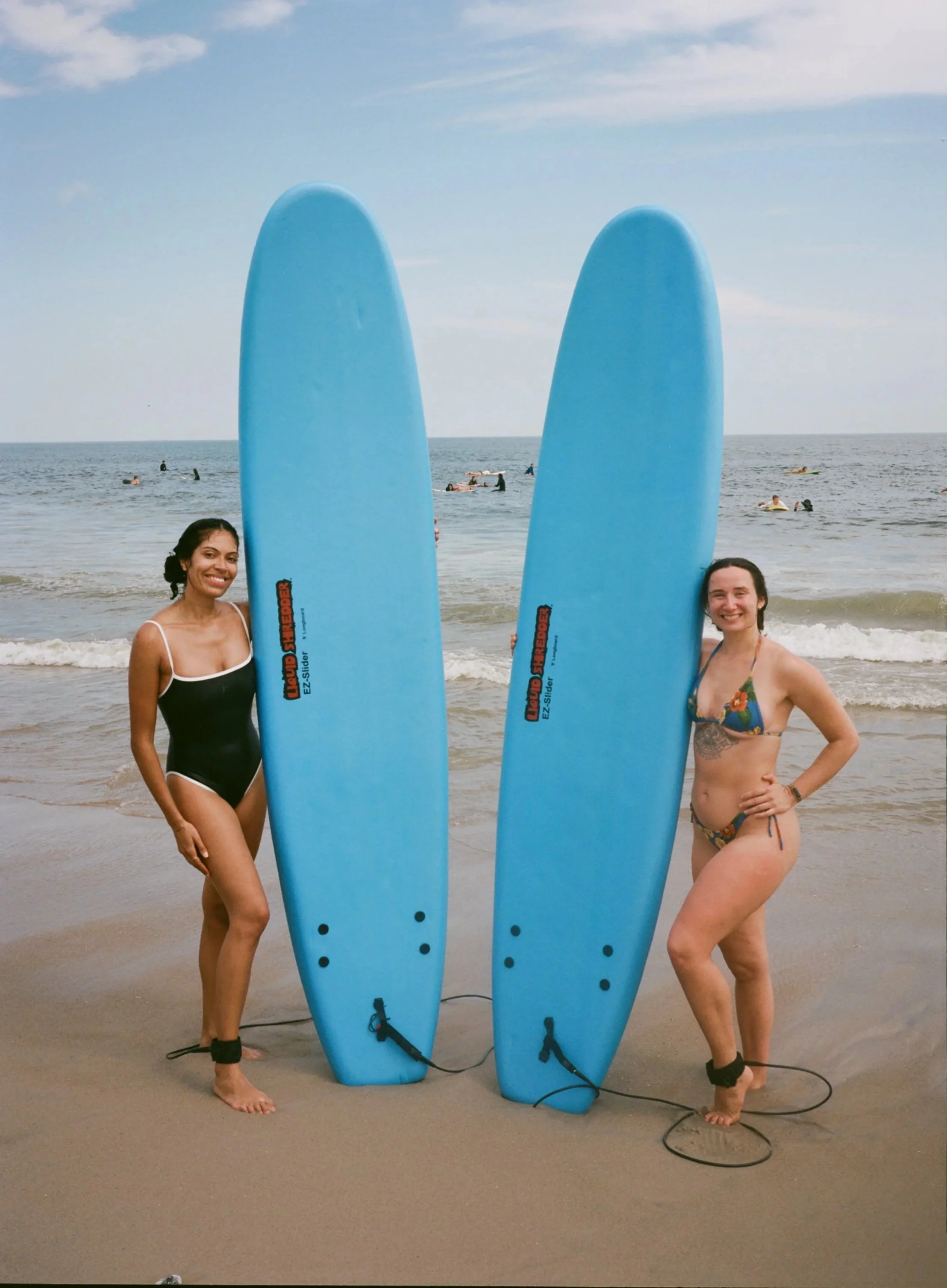 Two women in swimsuits standing on a beach holding large blue bodyboards with ankle straps.