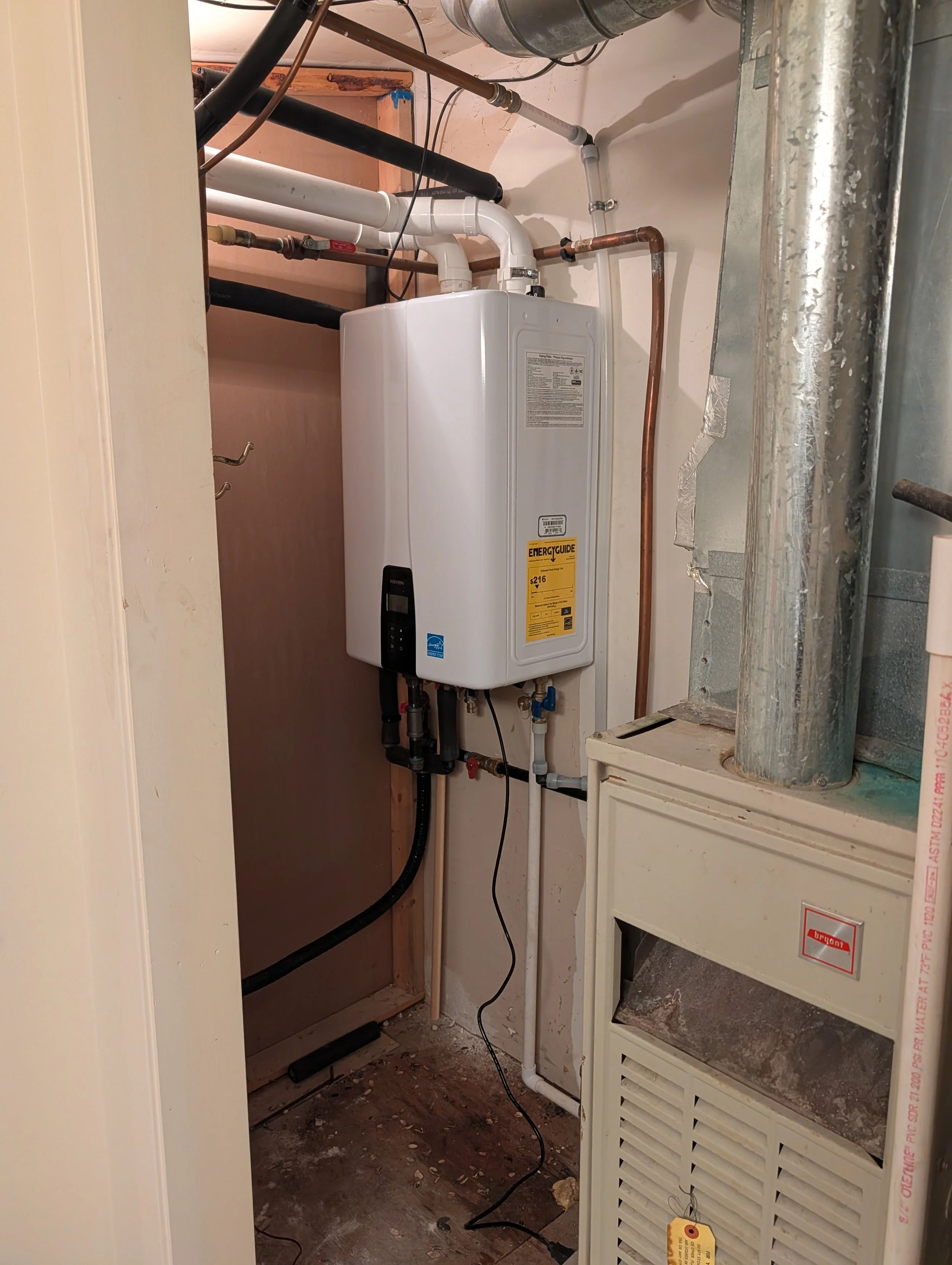 A utility closet with a water heater, copper and PVC pipes, and a furnace, partially enclosed with a drywall and wooden framing.