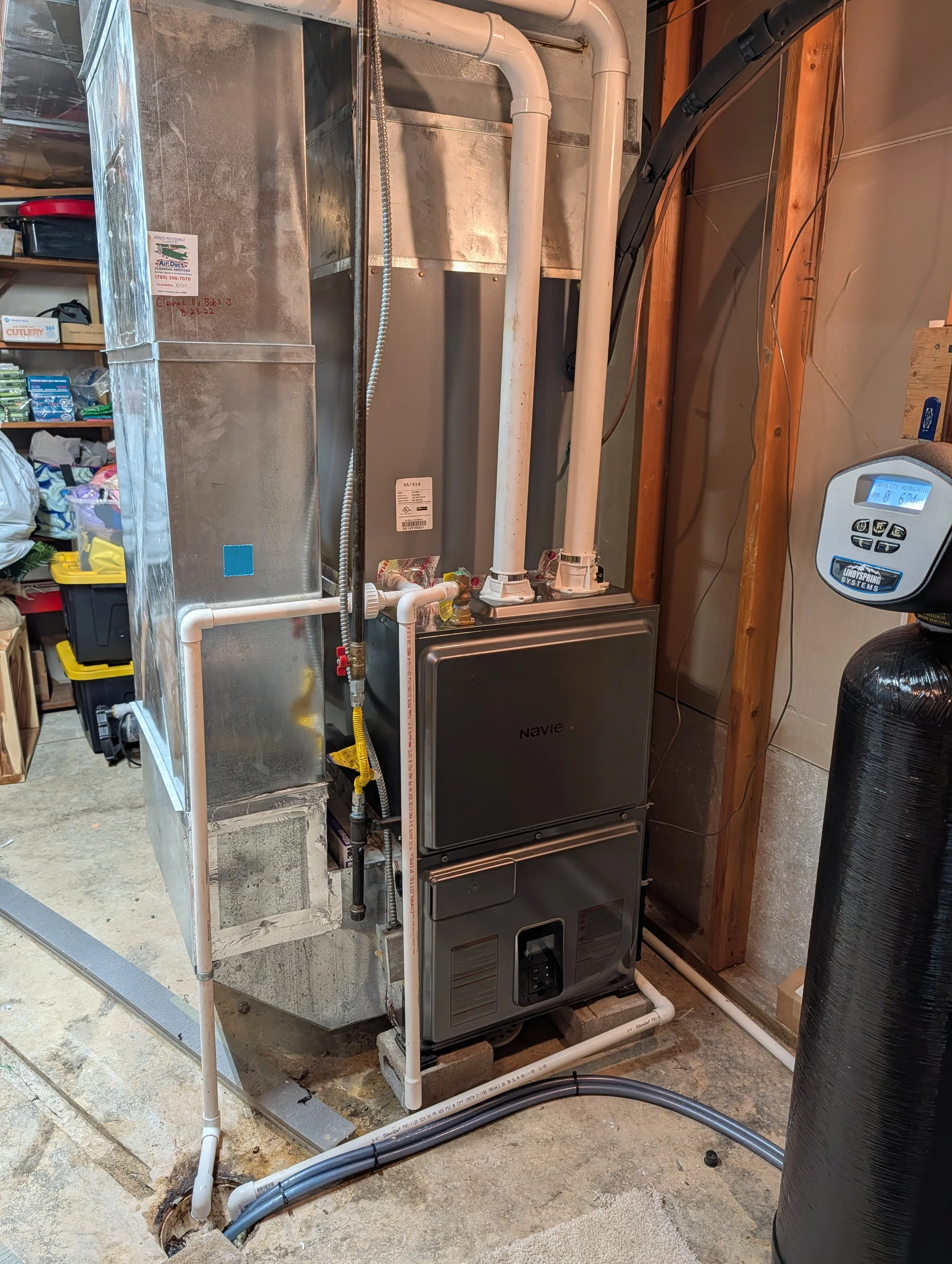 A furnace and ventilation pipes installed in a basement or utility room, with pipes running out of the top and side of the unit, and a black water softener tank to the right.
