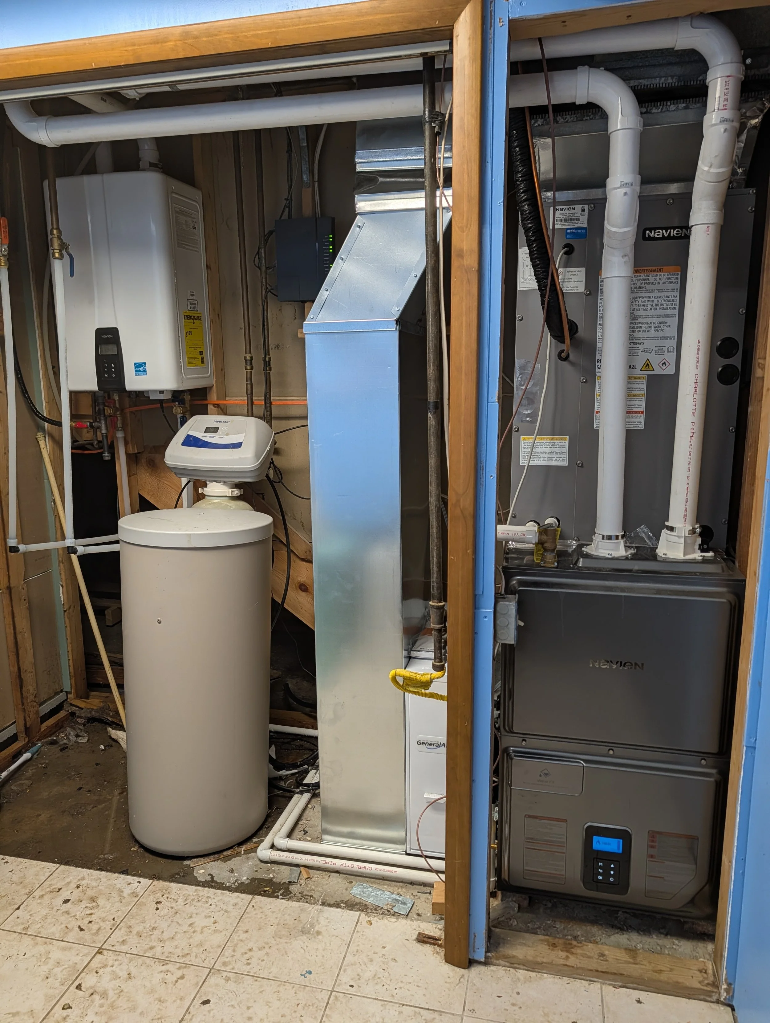 A utility room under construction with plumbing, HVAC units, and a water softener system installed on a wooden frame. The floor is tiled, and there are visible pipes and electrical components.