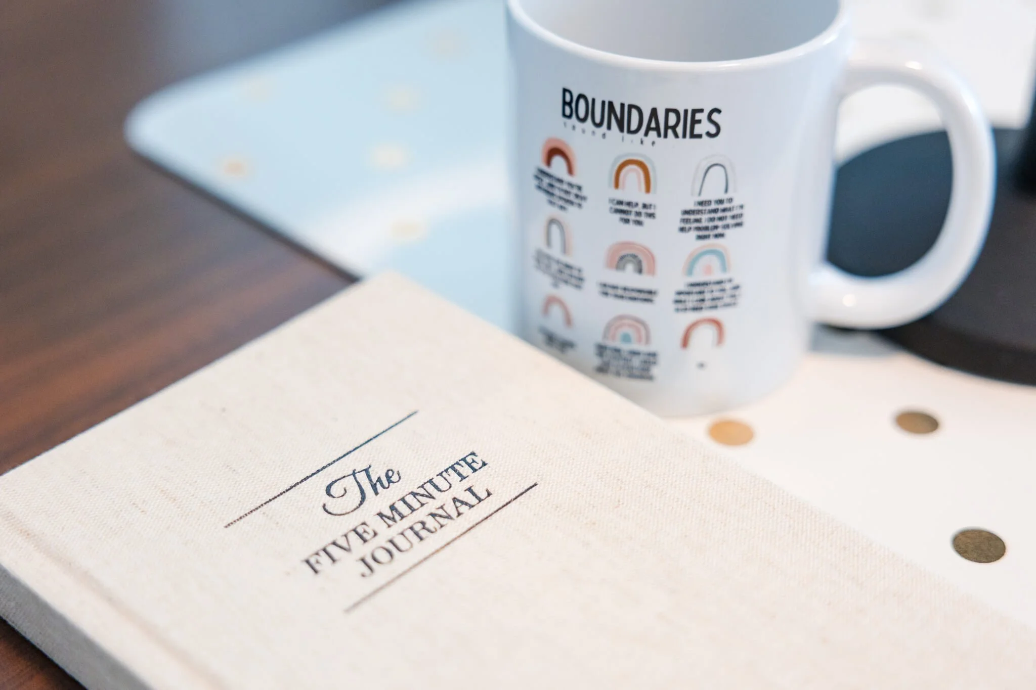 A white notebook titled 'The Five Minute Journal' is placed on a wooden surface, with a white mug labeled 'Boundaries' featuring various rainbow and line illustrations in the background.