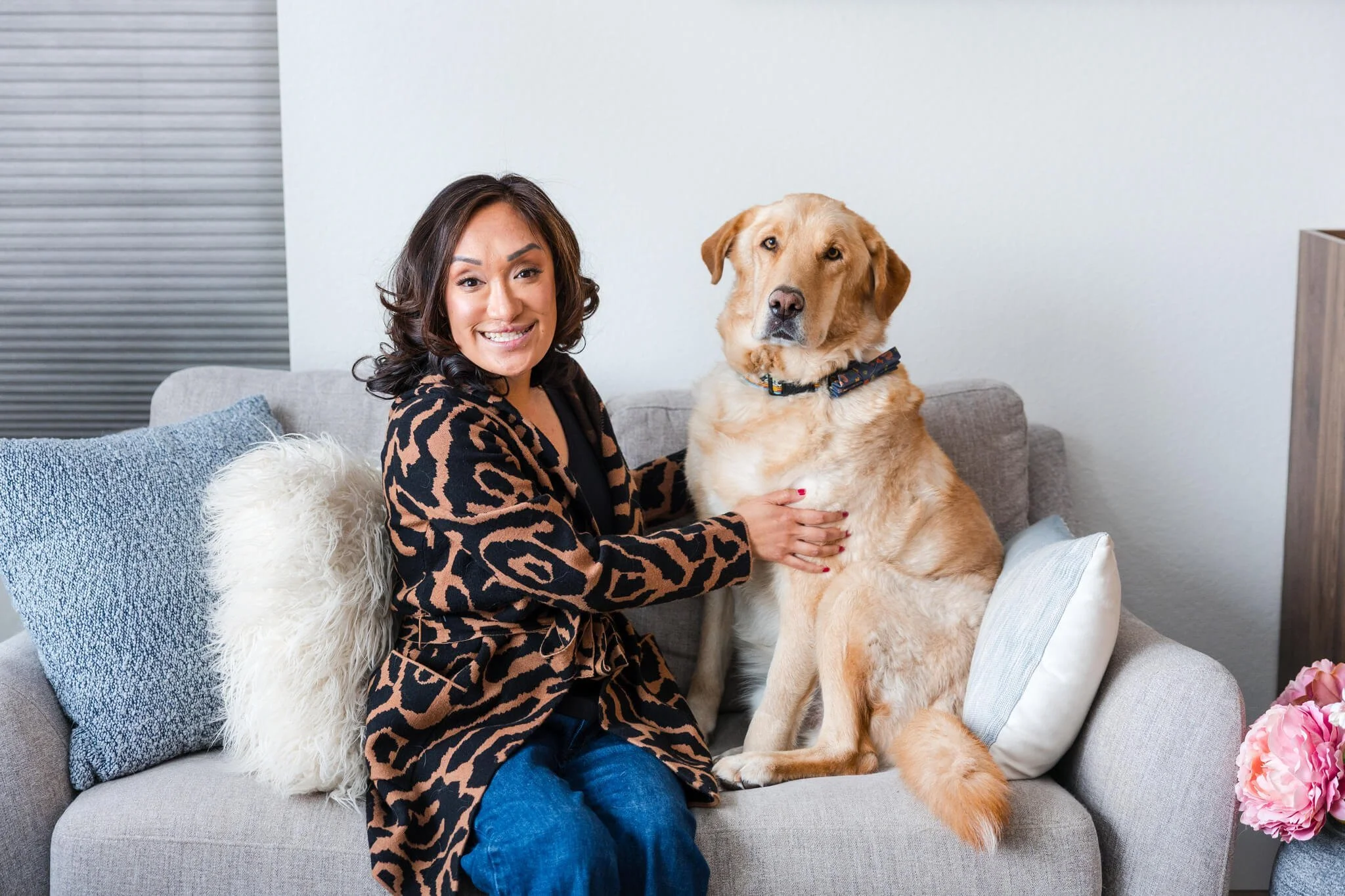 A woman with dark hair and a big smile, wearing a black and brown leopard print jacket, sitting on a gray sofa, holding a large yellow Labrador retriever dog with a collar, surrounded by blue, white, and furry pillows, in a cozy living room.