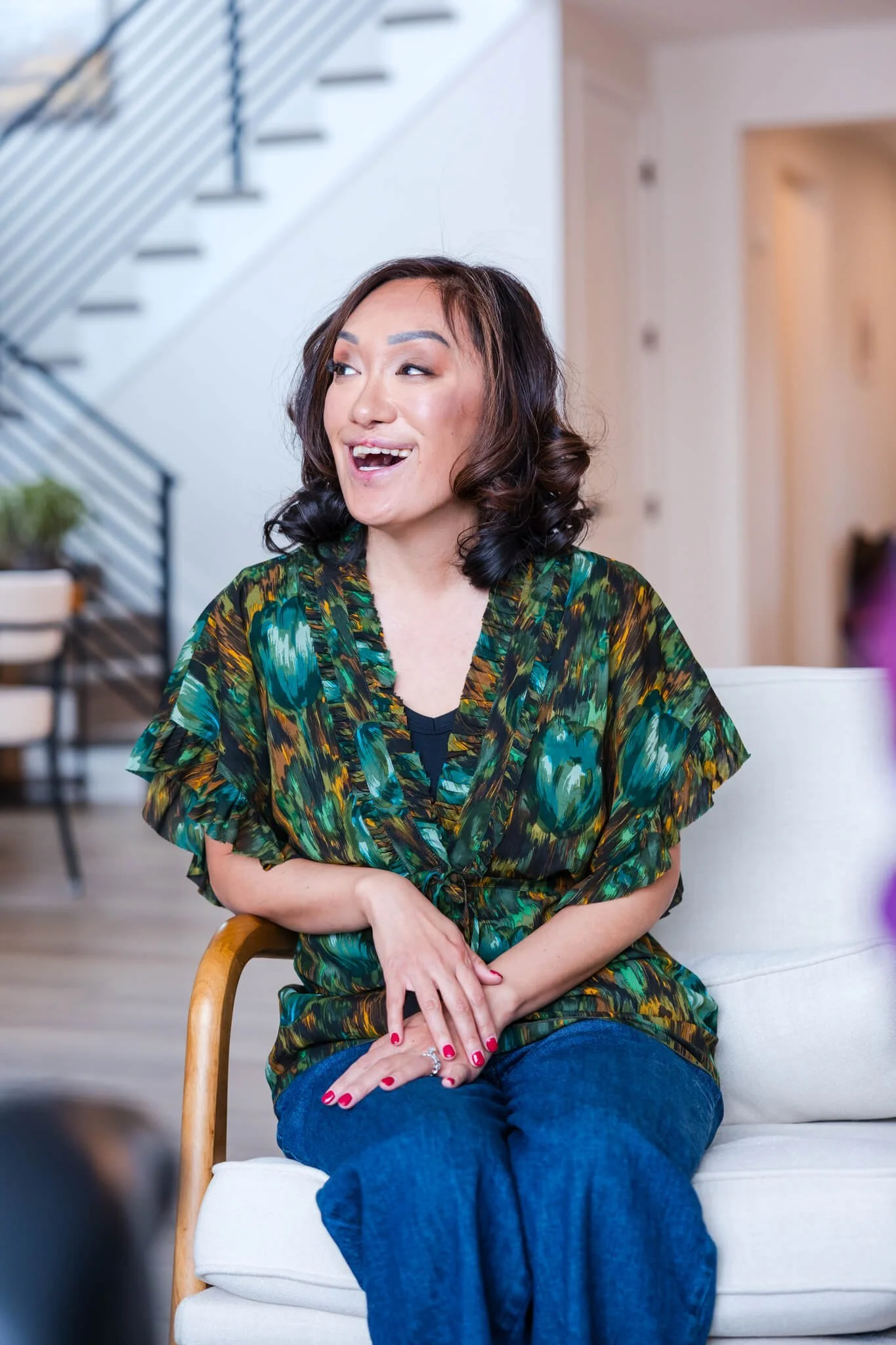 A woman with dark, wavy hair smiling and talking while seated on a white cushioned chair inside a home, wearing a colorful green and black patterned blouse and blue jeans.