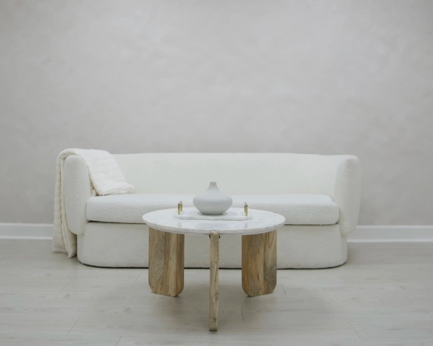 Minimalist living room with a white sofa, a white vase on a small round marble table, and a beige throw blanket. The room has light-colored wooden flooring and a plain light gray wall.
