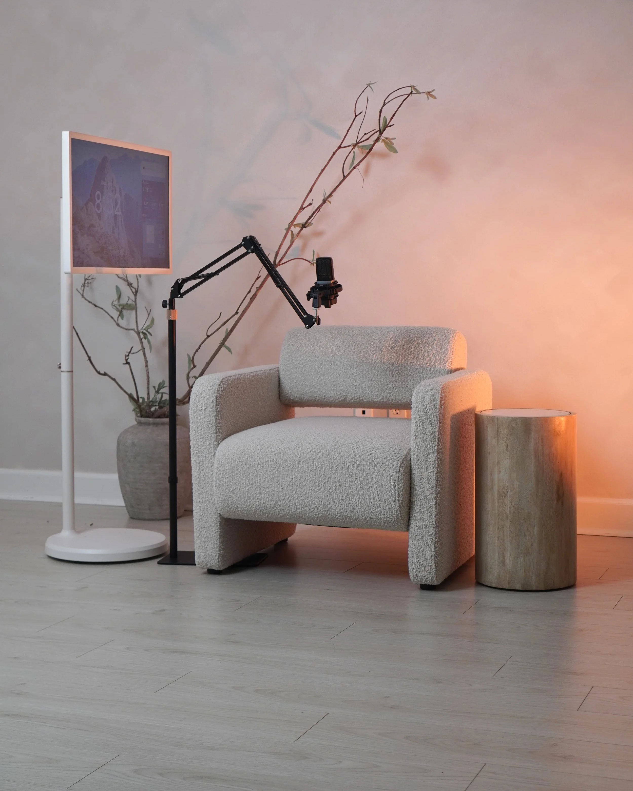 A cozy living space with a beige armchair, a wooden side table, a white floor lamp, a bed with a white textured cover, a potted plant with long branches, a microphone on an adjustable arm, and a wall displaying a computer monitor with a scenic backgr