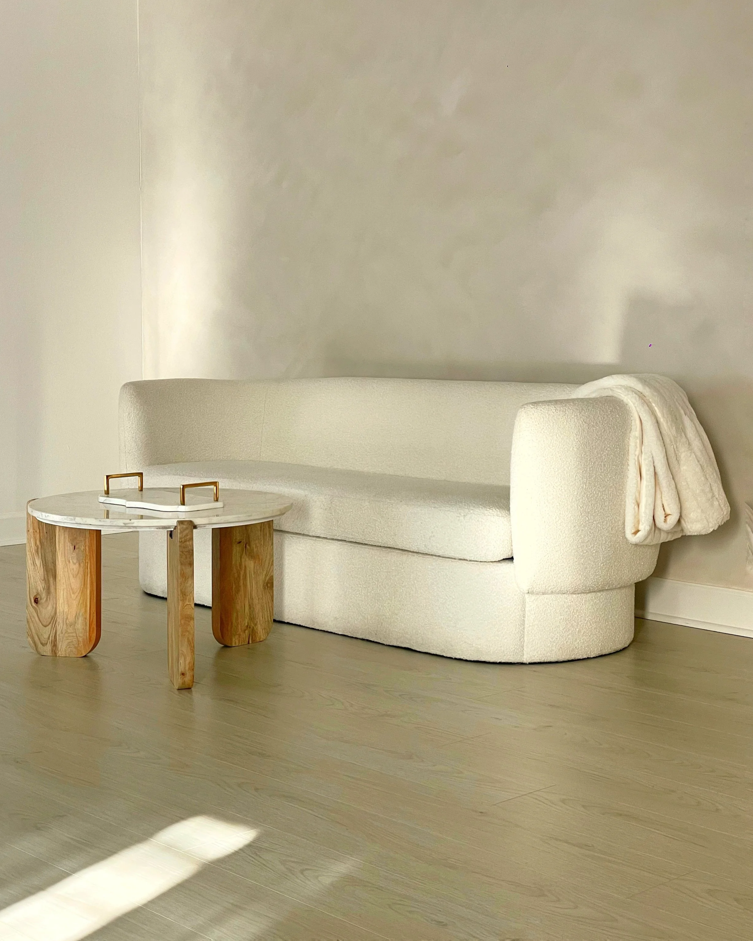 A cozy corner with a cream-colored curved sofa, a small round side table with a marble top and wooden legs, and a folded cream blanket resting on the sofa arm. The wall behind is plain and light-colored, and there is sunlight casting white streaks on