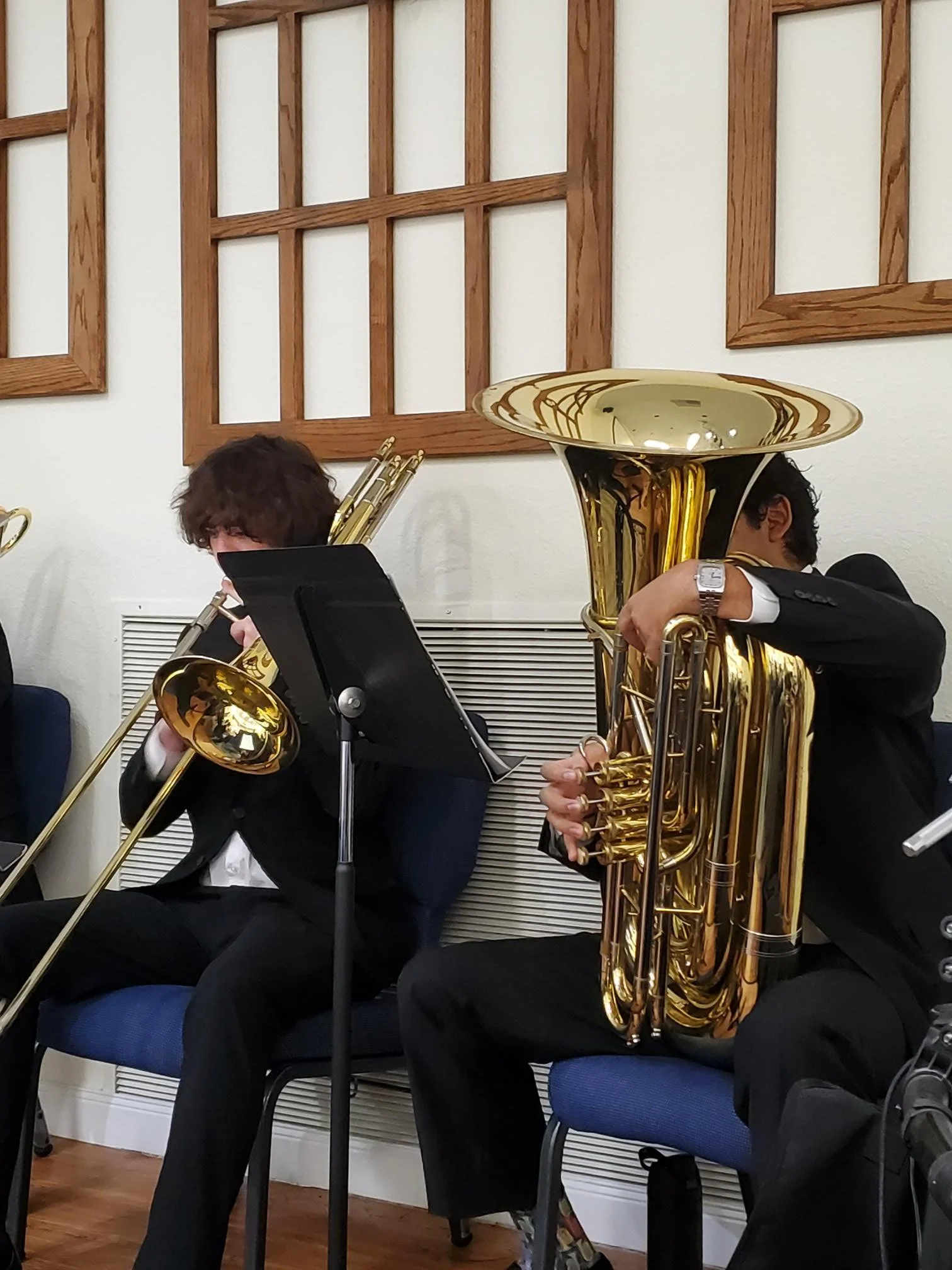 Musicians playing brass instruments during a performance, seated on chairs in a room with wooden wall decor.