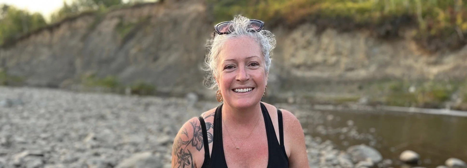 Smiling Loretta Ransom with curly gray hair, sunglasses on her head, and a tattoo on her shoulder, outdoors near a rocky riverbank.