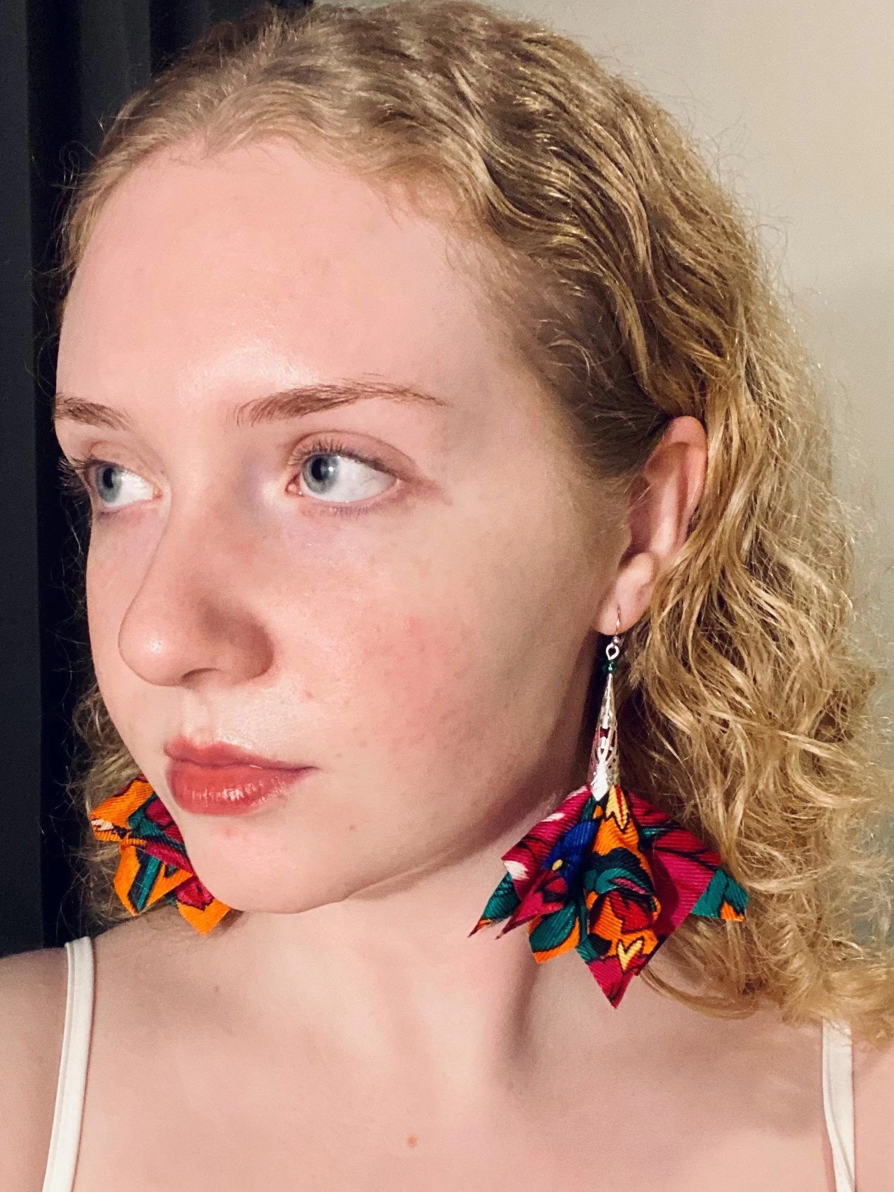 A woman with curly red hair wearing handcrafted colorful, patterned earrings and a white tank top.