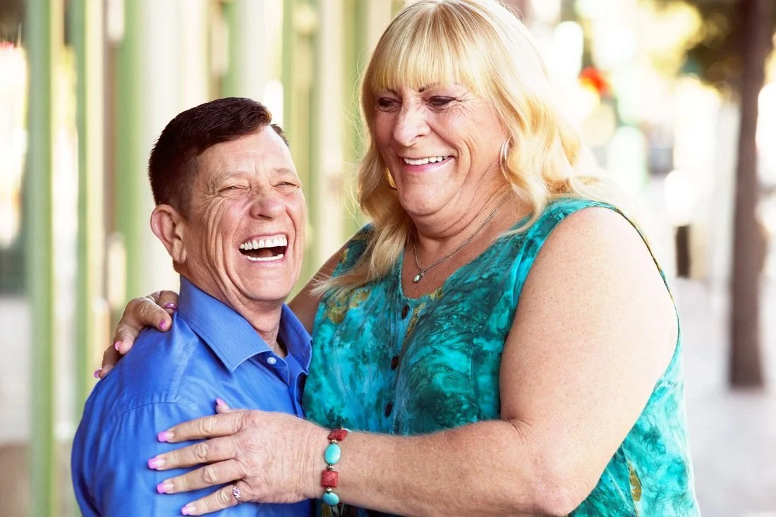Two people, one with longish blond hair and another with short brown hair happily hugging and laughing outdoors on a sunny day.
