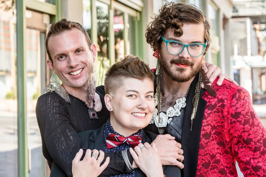 Three people posing closely together outdoors with urban background, dressed in vibrant and fashionable clothing.