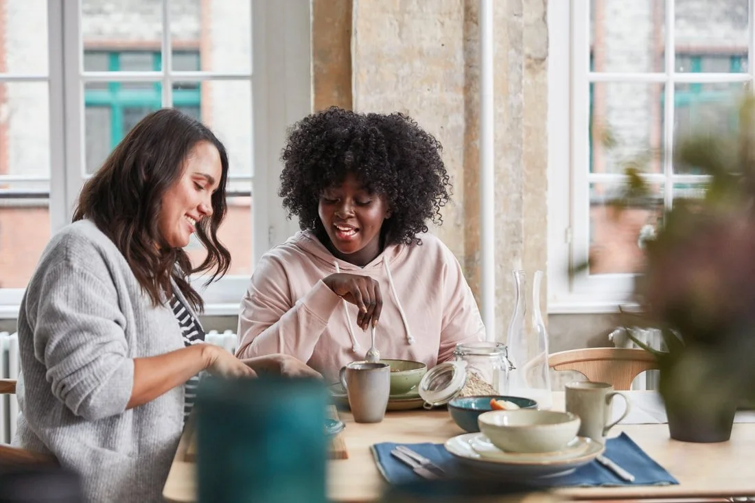 Two people, one with long straight brown hair and another with a shortish brown afro sitting at a table, smiling, having breakfast together, with dishes and cups on the table, near windows with a view of buildings outside.