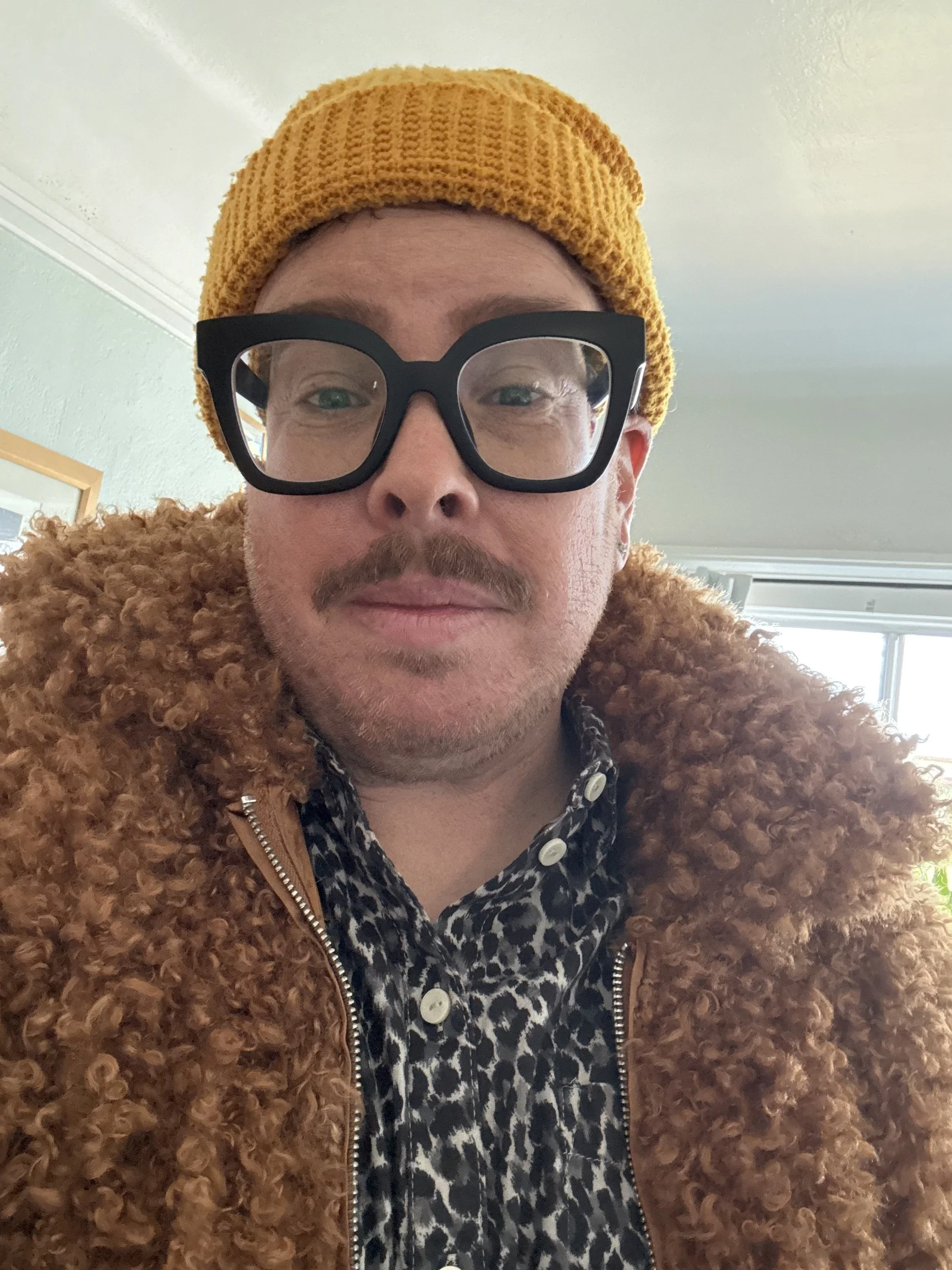 A person with light skin and a large brown moustache wearing a mustard yellow knit beanie, large black glasses with clear lenses, a patterned shirt, and a brown curly fur coat.
