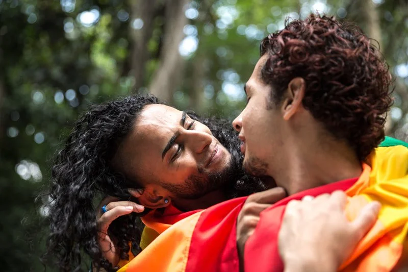 Two people embracing outdoors, with the person on the left smiling and looking into the eyes of the person on the right, surrounded by trees and natural light.
