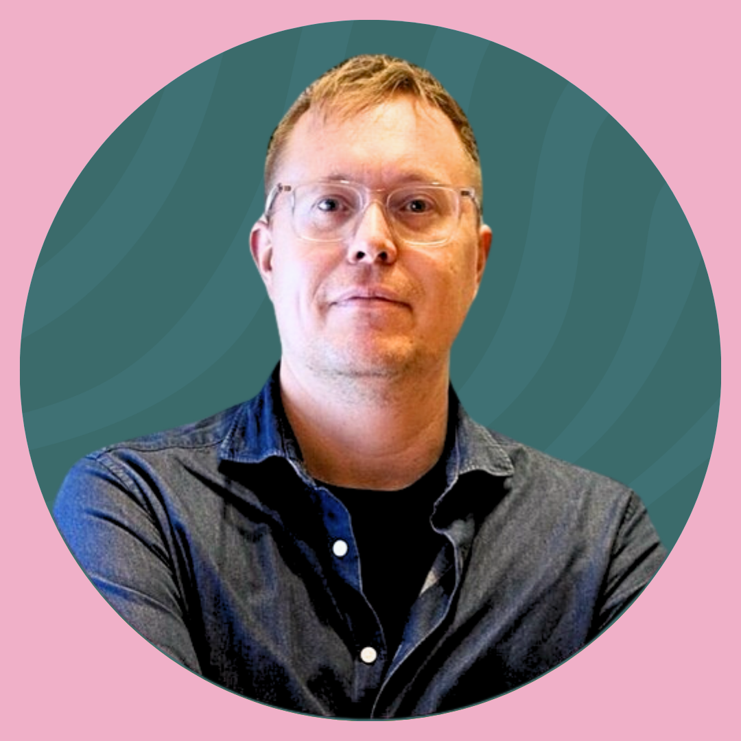 A man in a gray suit, white shirt, wearing glasses, smiling outdoors with green foliage in the background.