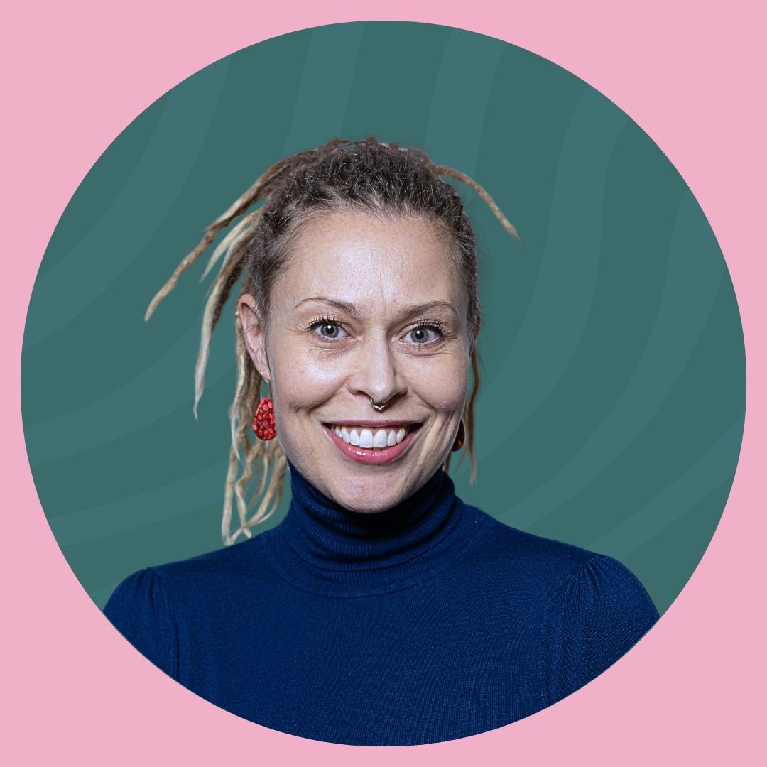 Close-up of a smiling woman with blonde dreadlocks, some with beads, and a septum piercing, standing in front of a brick wall.
