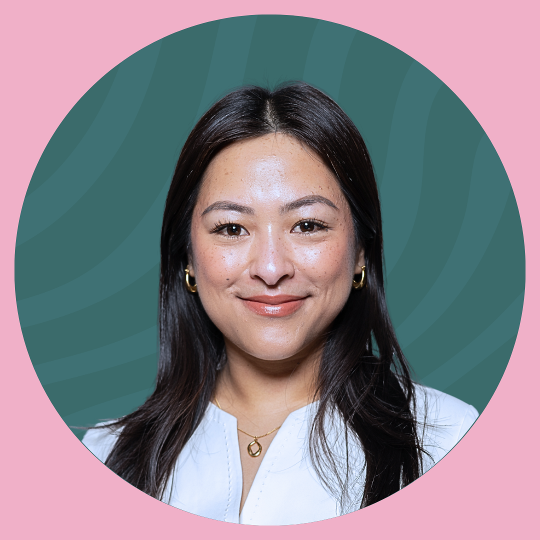 A woman with long black hair smiling at the camera inside a lush greenhouse with green plants and a small water feature.