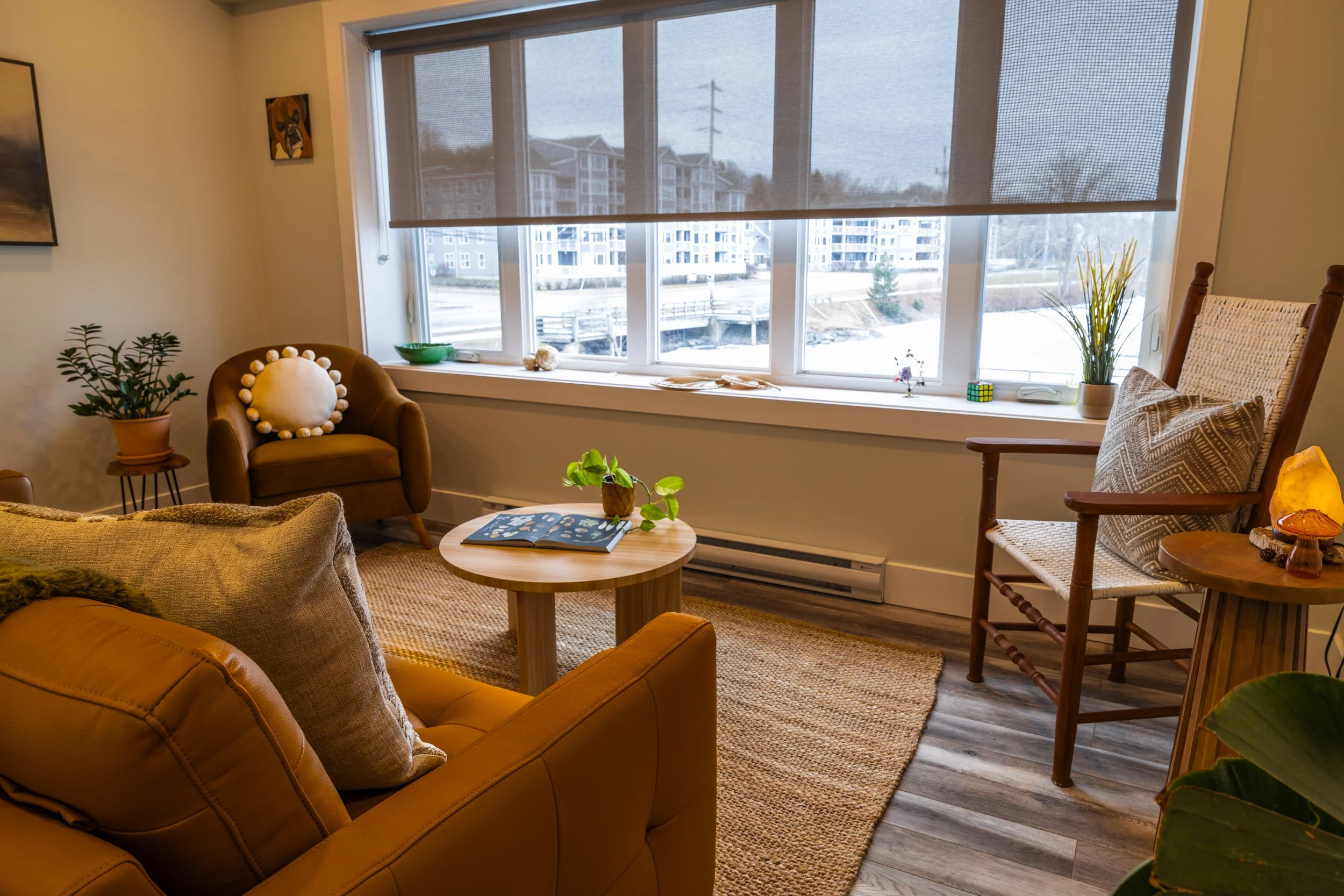A cozy living room with large windows showing a snowy outdoor scene and modern apartment buildings. The room has a tan armchair with a round cushion, a small round wooden coffee table with an open book and a potted plant, other chairs with decorative pillows, a tall potted plant, and artwork on the walls. The floor is covered with a woven rug.