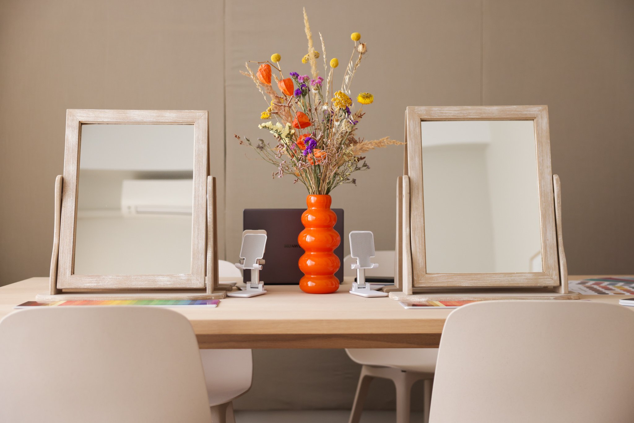 Two mirrors on a table with an orange vase containing colorful dried flowers between them, surrounded by white chairs and digital devices.