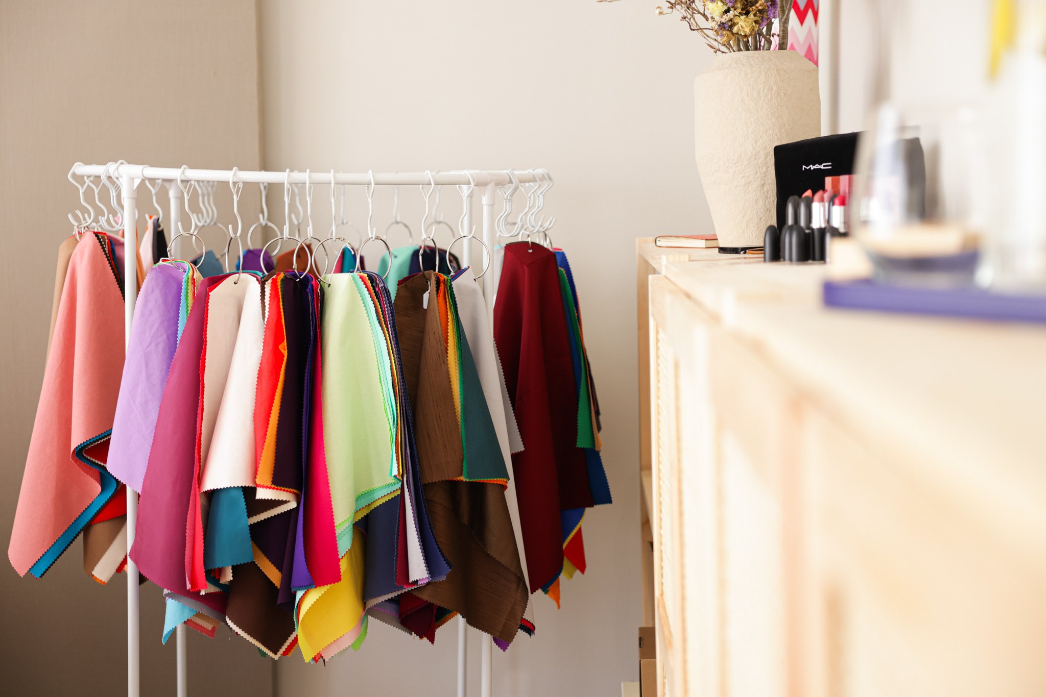 Colorful fabric swatches hanging on a rack next to a wooden counter with makeup items and a vase.