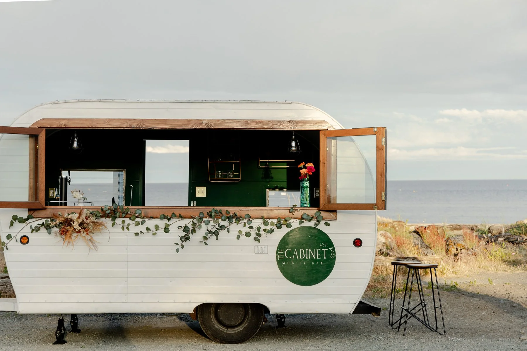 Vintage 1963 trailer bar rental in Victoria, BC, styled with greenery for an oceanfront wedding ceremony.
