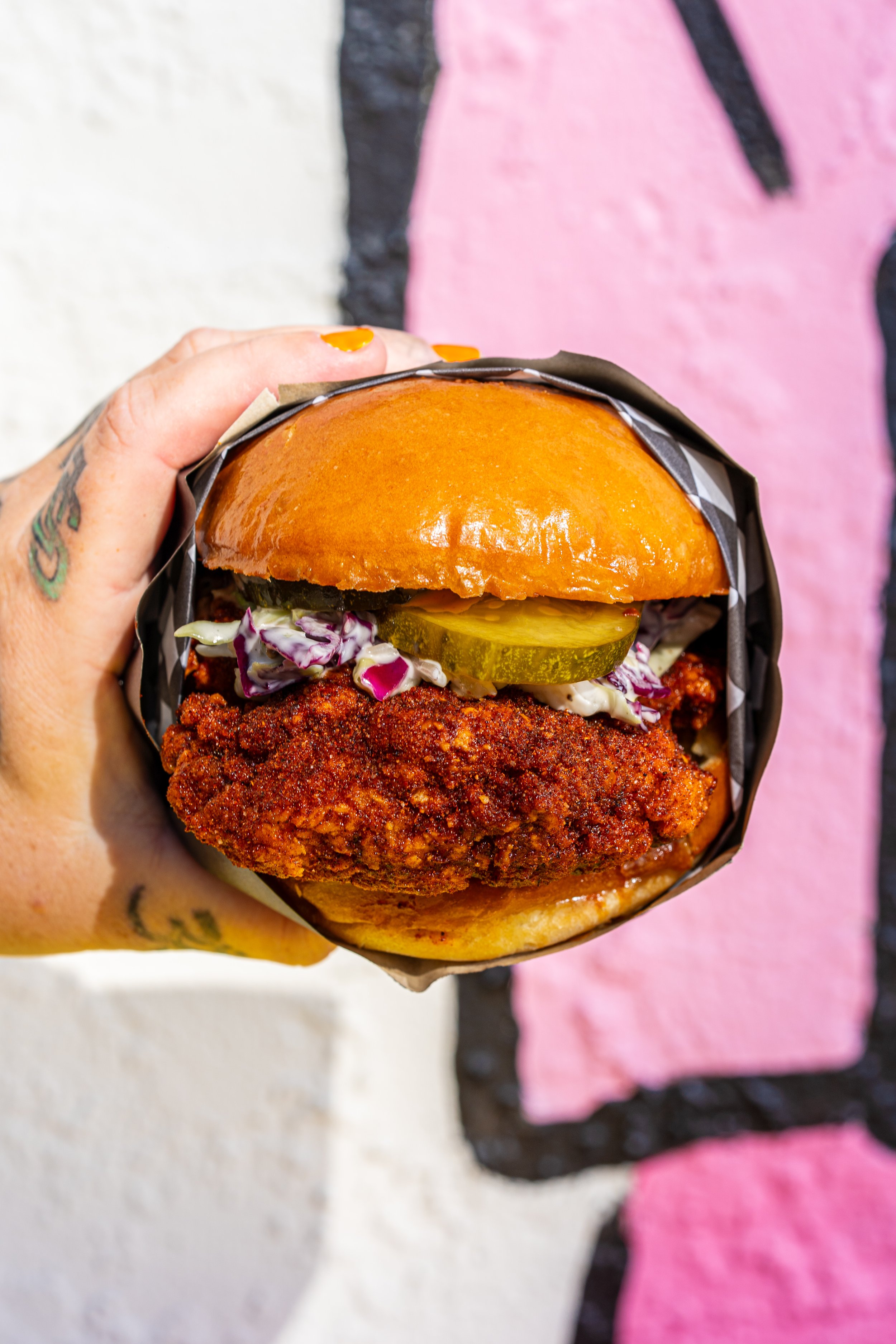 Close-up of a hand holding a fried chicken sandwich with pickles, lettuce, and sauce in a bun against a colorful graffiti background.