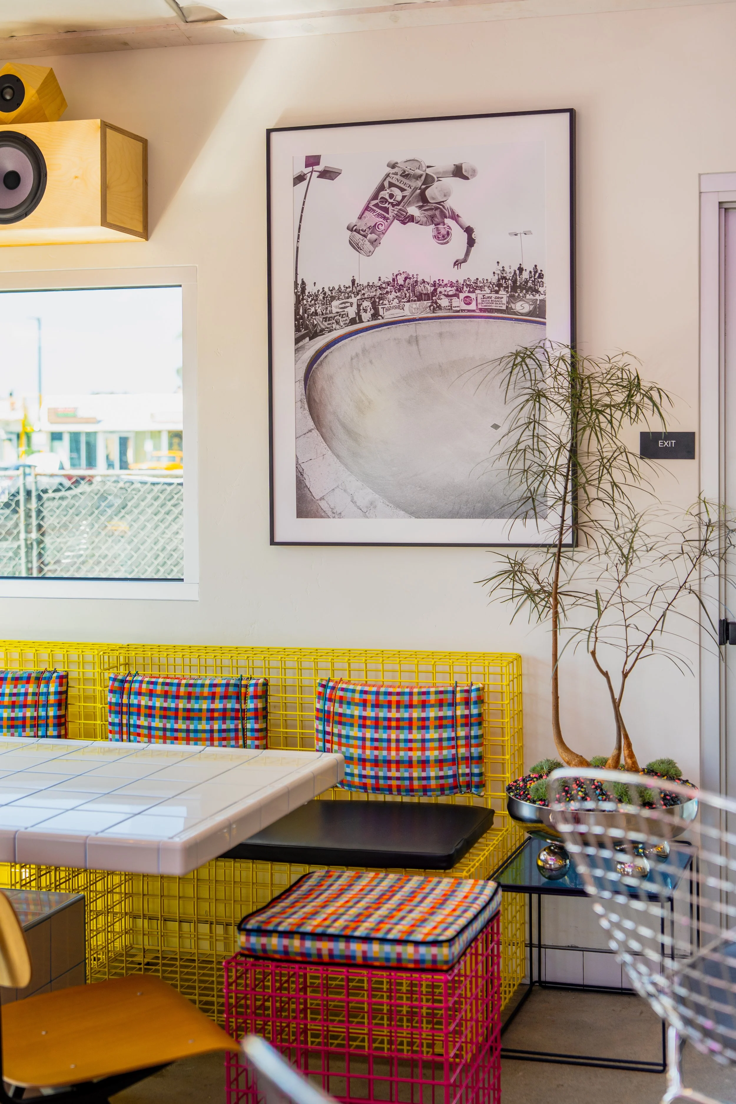 Interior of a colorful cafe with a white tiled table, patterned chairs, a yellow wire bench, and a skateboard-themed artwork on the wall.