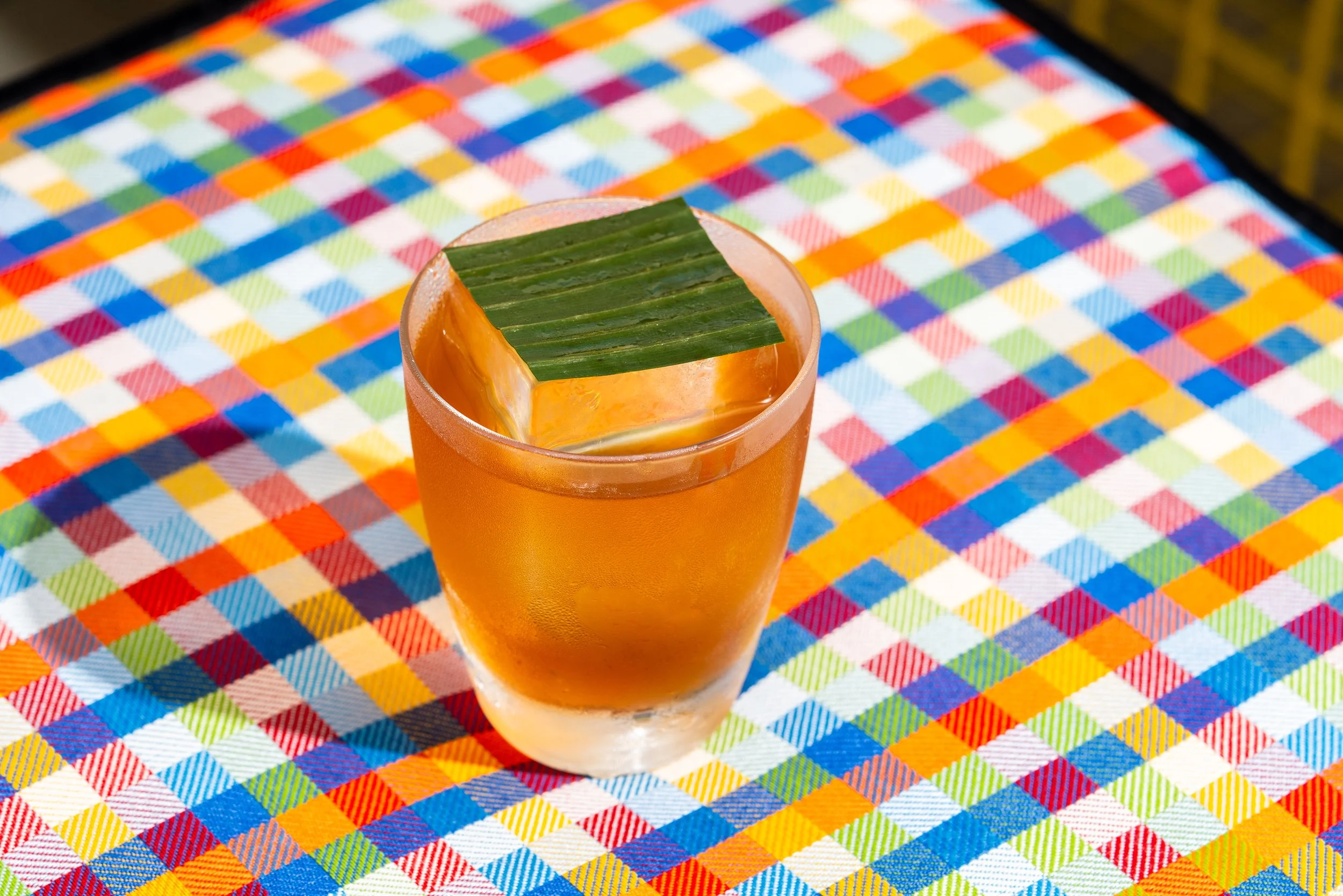 A glass of orange-colored beverage with a green leaf on top, placed on a colorful checkered tablecloth.
