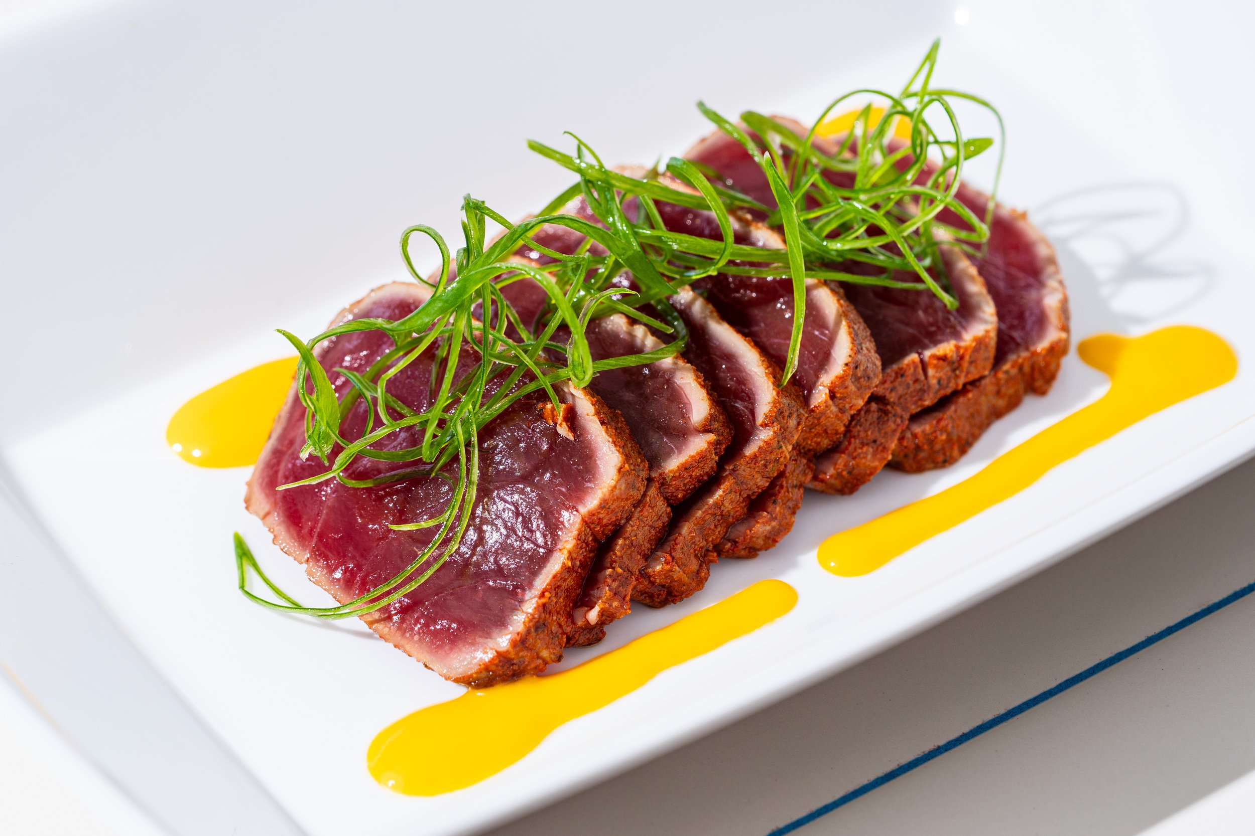Slices of seared tuna topped with green garnishes, drizzled with yellow sauce, on a white rectangular plate.