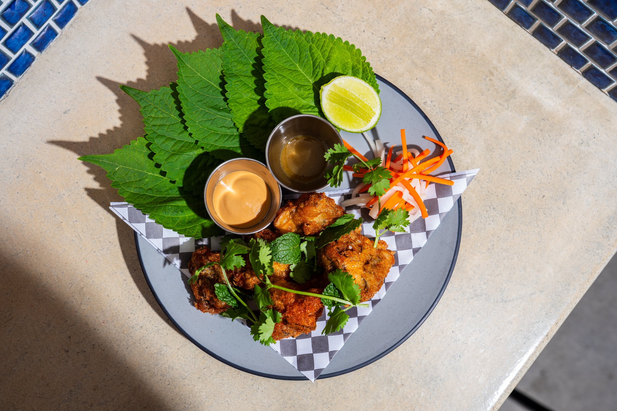 Plate of fried chicken pieces garnished with cilantro and fresh green leaves, served with two dipping sauces, shredded carrots and daikon radish, a lime wedge, and fresh mint and cilantro leaves on a checkered paper liner.