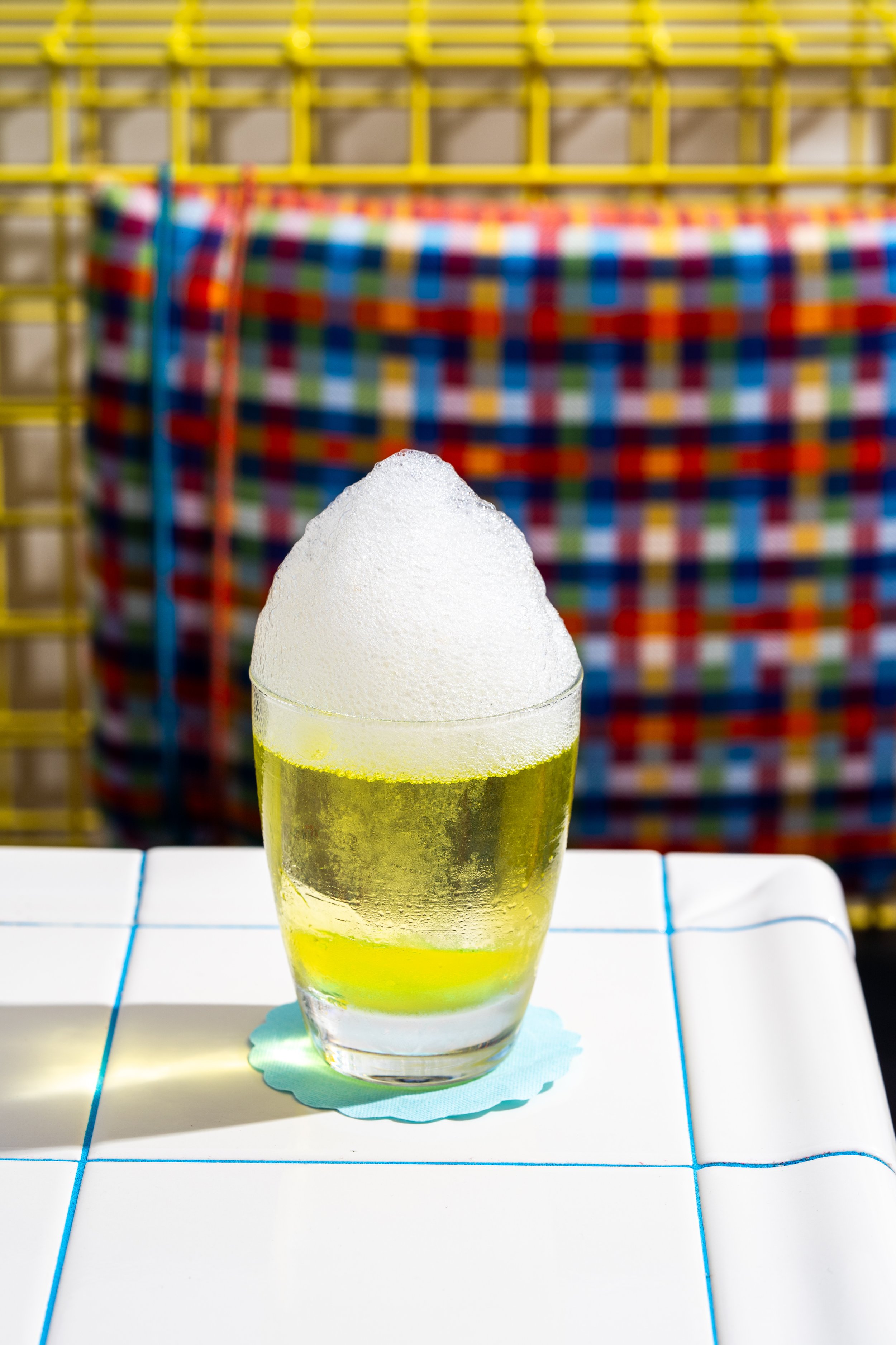 A glass of beer with foam on top placed on a tiled white table, with colorful fabric and yellow plastic grid in the background.