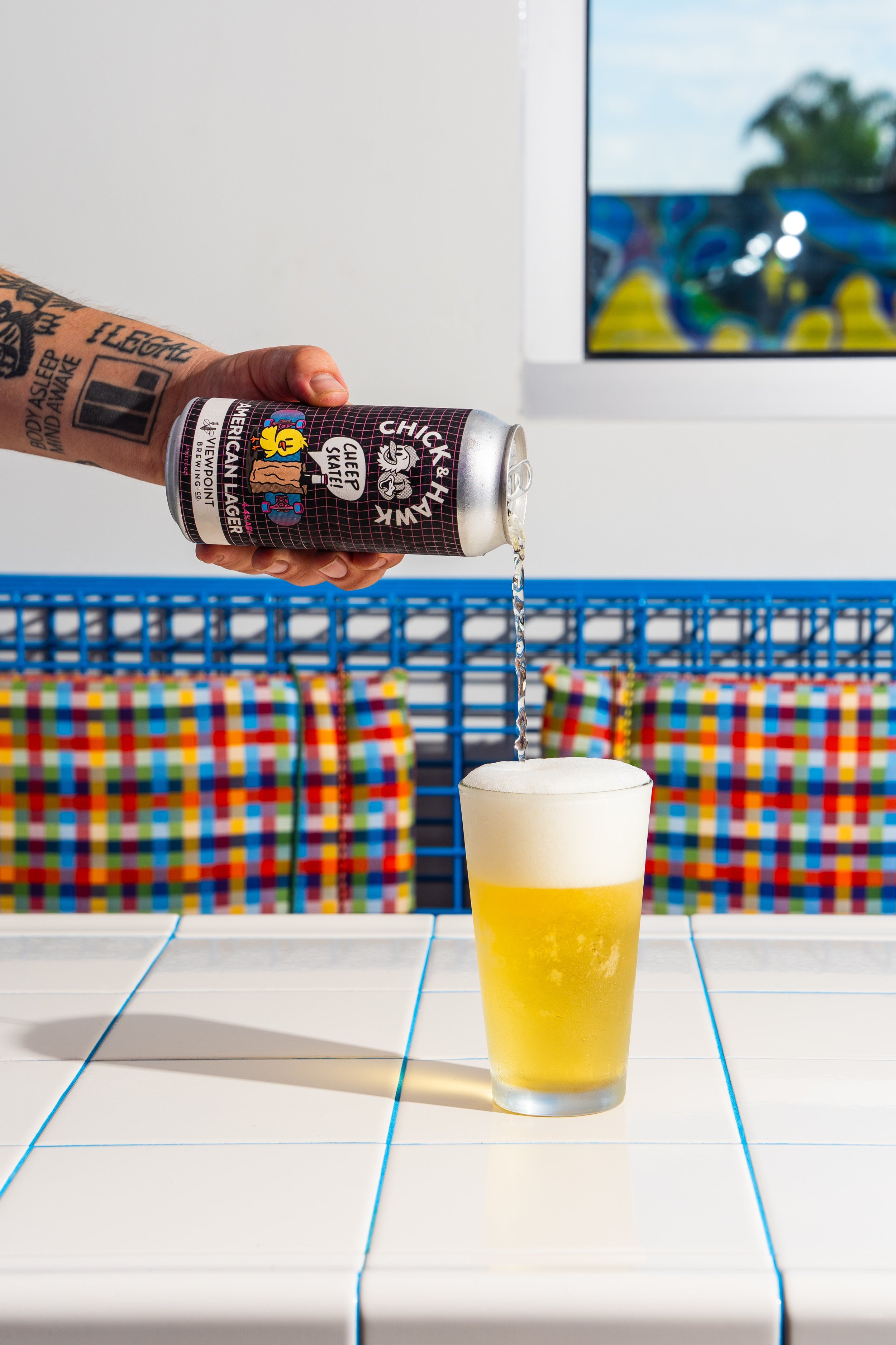 A hand holding a can of Chief & Hunk beer pouring beer into a glass with a foamy head on a white tiled surface.