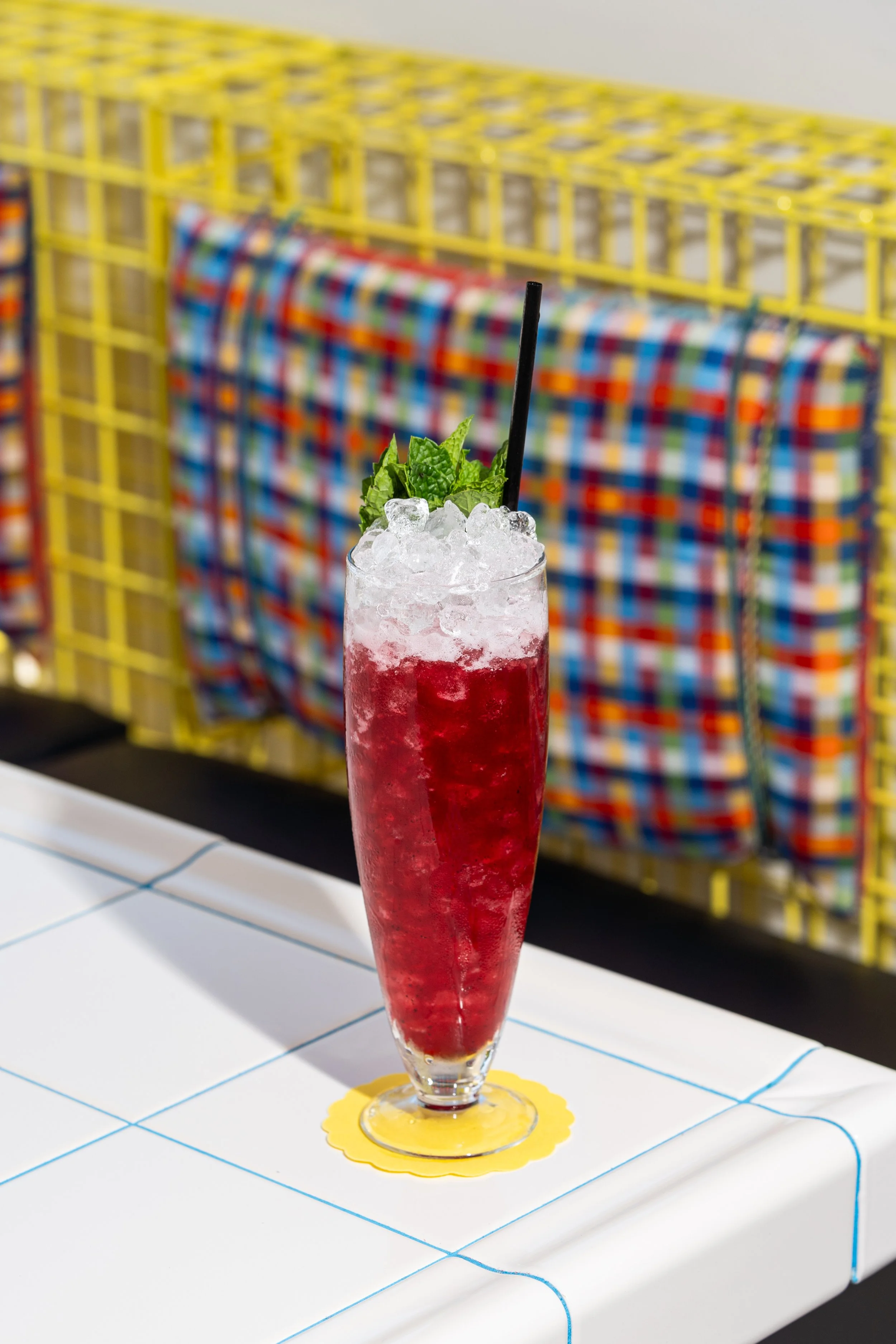 Red cocktail garnished with mint leaves and ice in a tall glass with a black straw, on a table with a yellow coaster.