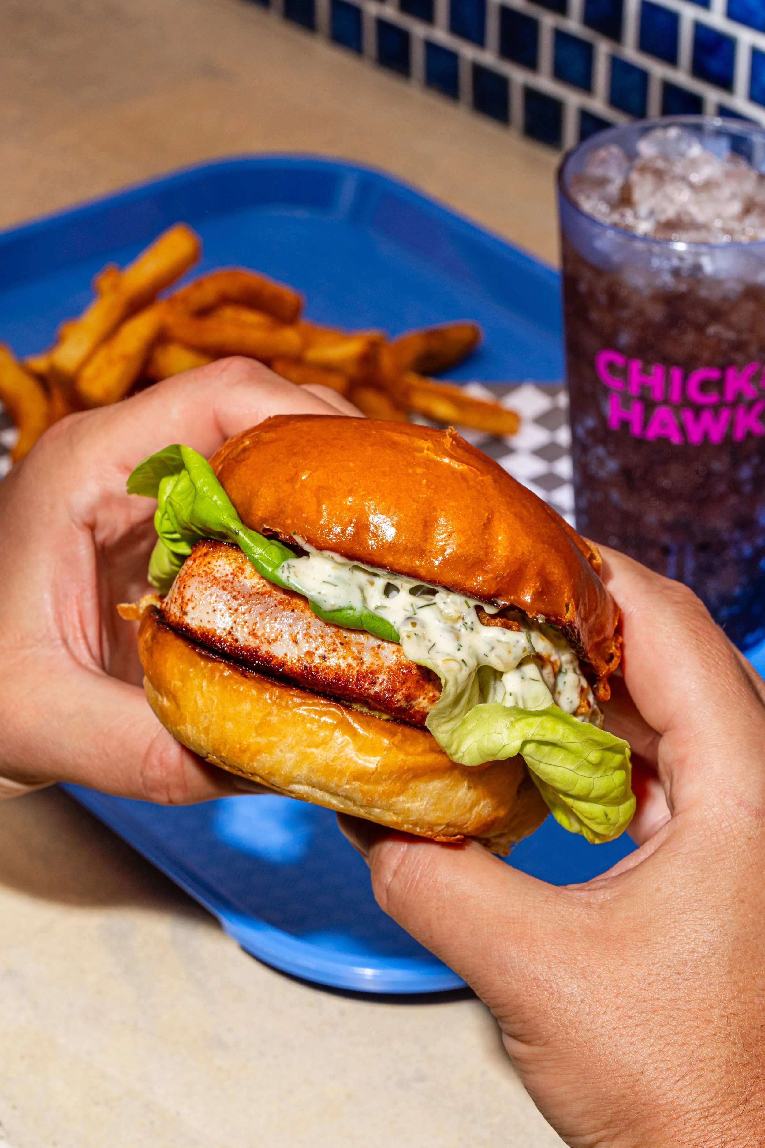 Person holding a sandwich with lettuce, seasoned fried fish, and creamy sauce in a bun, with French fries and a soda on a tray in the background.