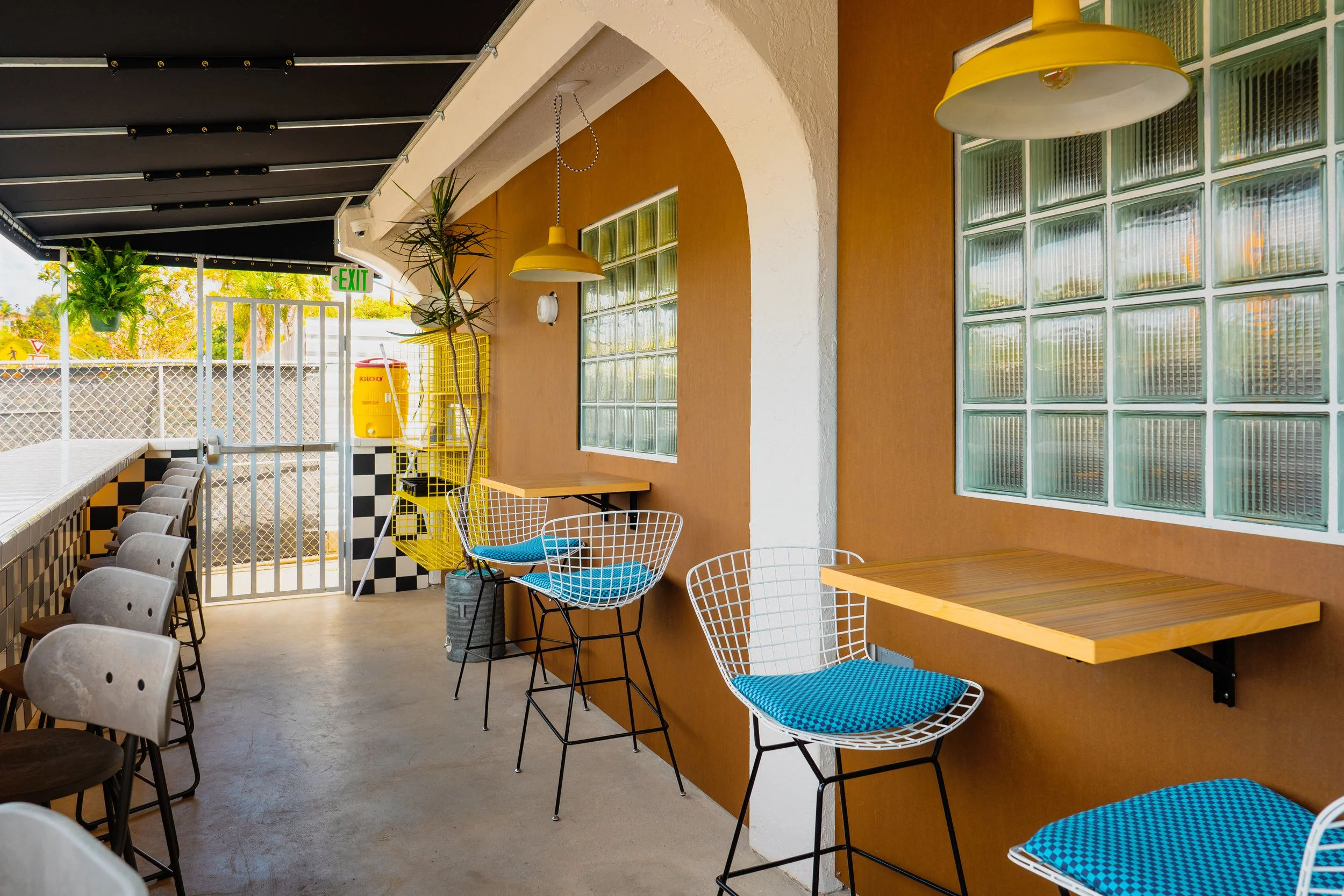 Interior of a modern cafe with wooden and metal furniture, yellow pendant lights, potted plants, glass block windows, and an outdoor seating area with a checkered wall and an exit door.